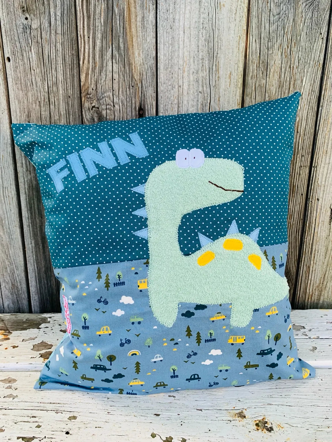 Handgenähtes Kinderkissen personalisiert, gestaltet petrol mit weißen Punkten im oberen Teil, rauchblau mit mit Autoprint im unteren Teil. Auf dem Kissen ist eine Dino-Applikation in mintgrünem Frottee.Fotografiert vor einem rustikalen Holzhintergrund