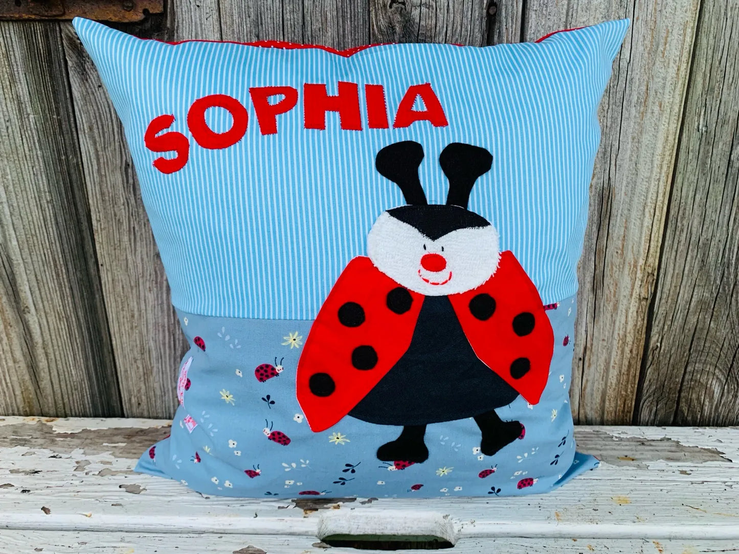 Handgenähtes Kinderkissen personalisiert, gestaltet hellblau mit weißen Streifen im oberen Teil,
rauchblau mit Marienkäferprint im unteren Teil. Auf dem Kissen ist eine Marienkäfer-Applikation in leuchtendem Rot.Fotografiert vor einem rustikalen Holzhintergrund