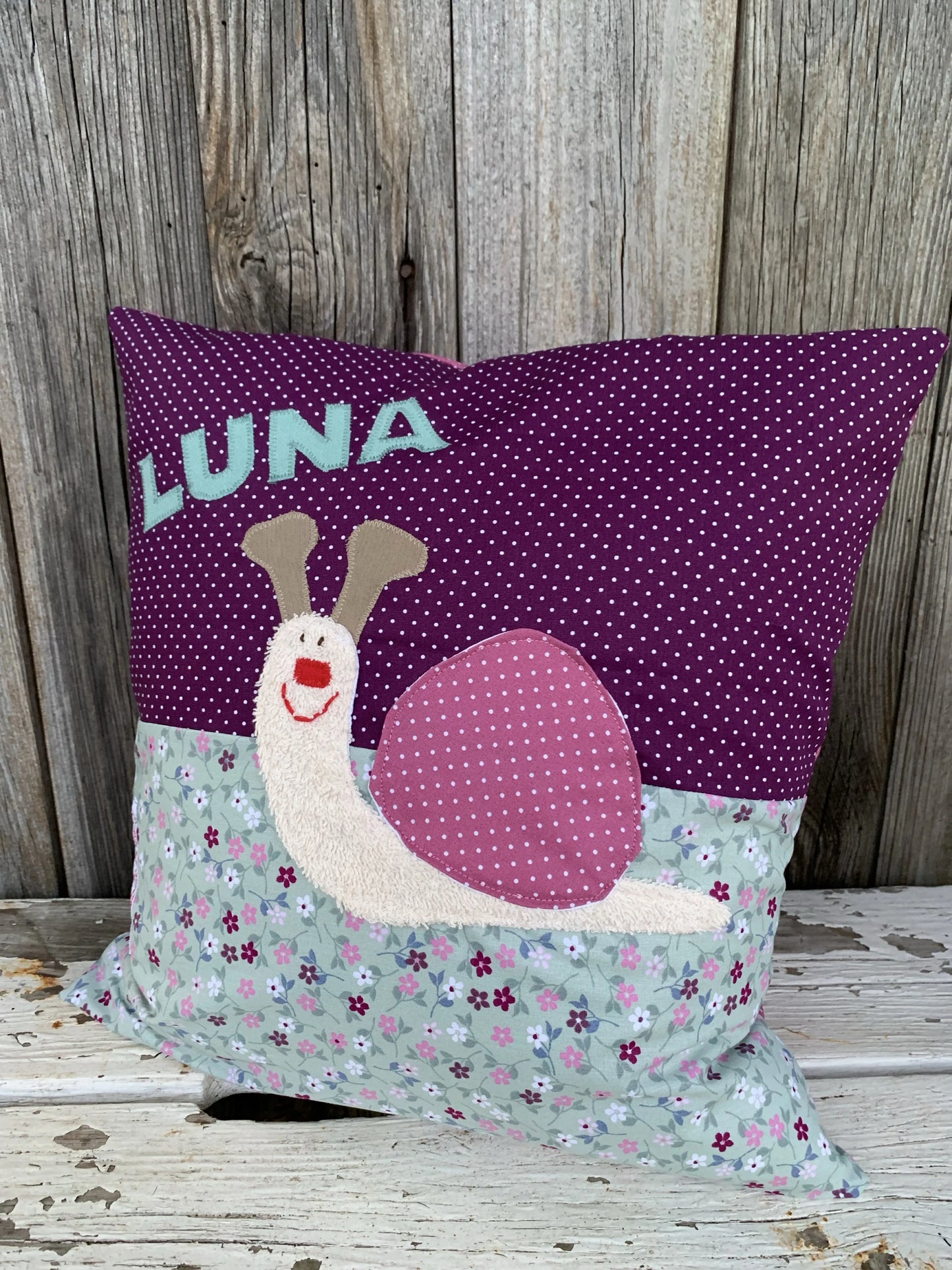 Handgenähtes Kinderkissen personalisiert, gestaltet dunkellila mit weißen Punkten im oberen Teil, altmint mit bunten Blümchen im unteren Teil. Auf dem Kissen ist eine
Schnecken-Applikation.Fotografiert vor einem rustikalen Holzhintergrund