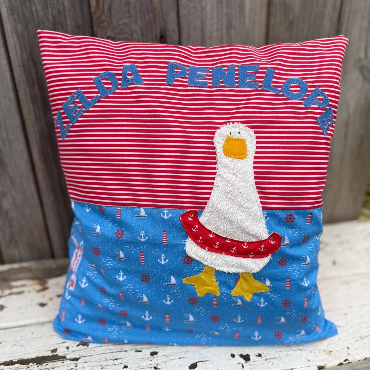 Handgenähtes Kinderkissen personalisiert, gestaltet rot mit weißen Streifen im oberen Teil, blau im maritimen Design im unteren Teil. Auf dem Kissen ist eine Enten-Applikation mit Schwimmreifen in weißem Frottee. Fotografiert vor einem rustikalen Holzhintergrund