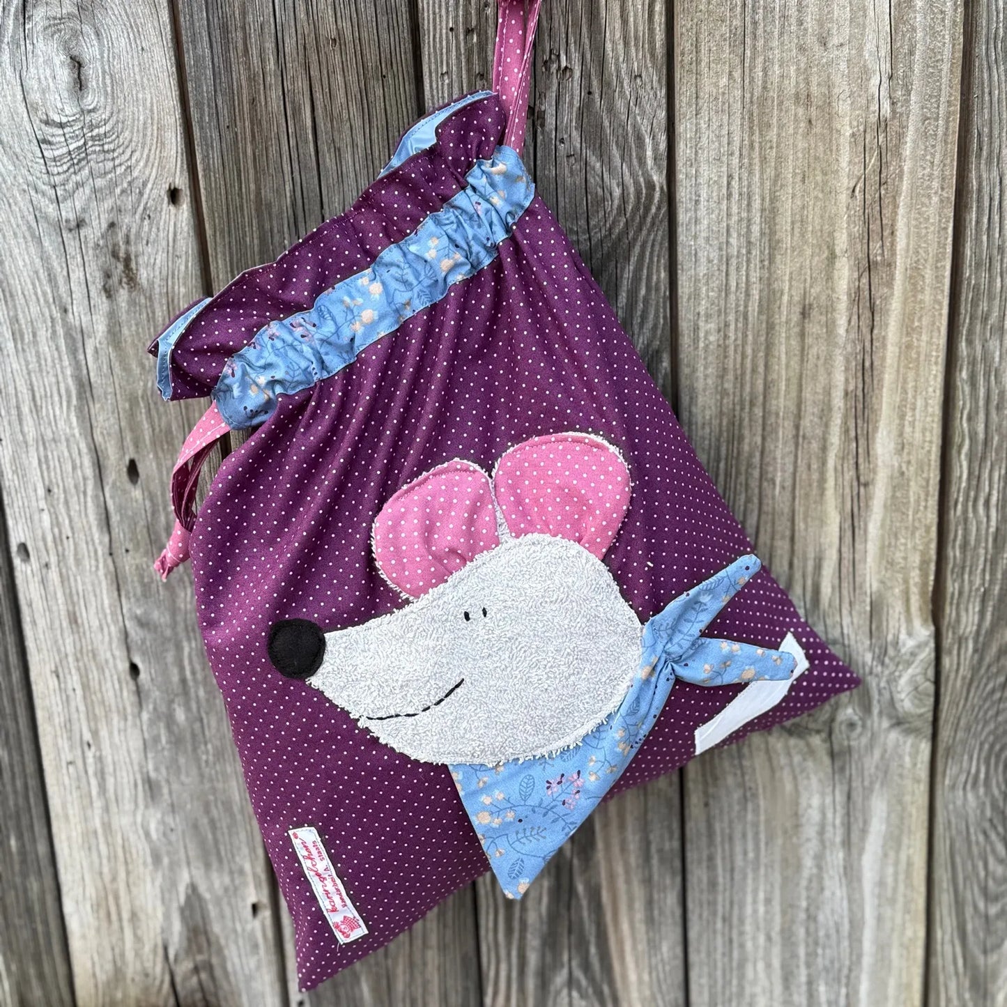 Hellgrüner Wetbag mit weißen Punkten, verziert mit einer bunten Stoffapplikation einer Maus aus grauem Frottee .DerBeutel hat einen floralen Tunnelzug und altrosa Kordeln, aufgenommen vor rustikalem Holzhintergrund.
