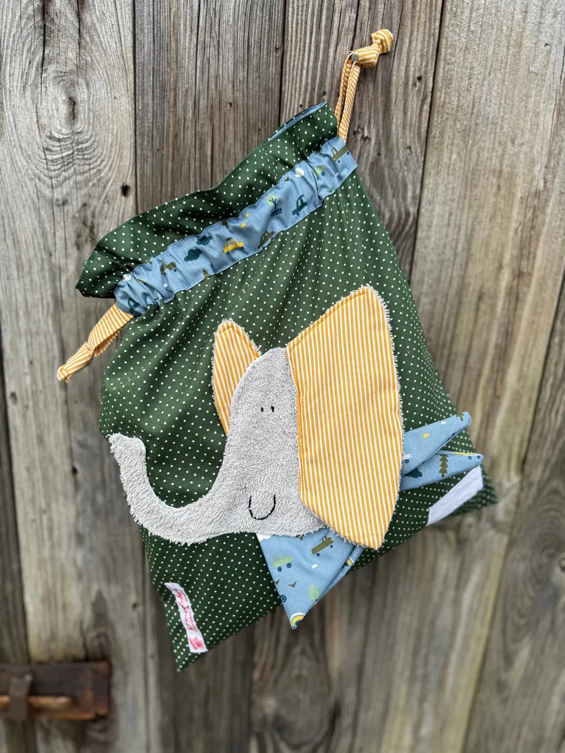 Dunkelgrüner Wetbag mit weißen Punkten, verziert mit einer bunten Stoffapplikation einem Elefant .DerBeutel hat einen blauen Tunnelzug aus und senfgelben Kordeln, aufgenommen vor rustikalem Holzhintergrund.