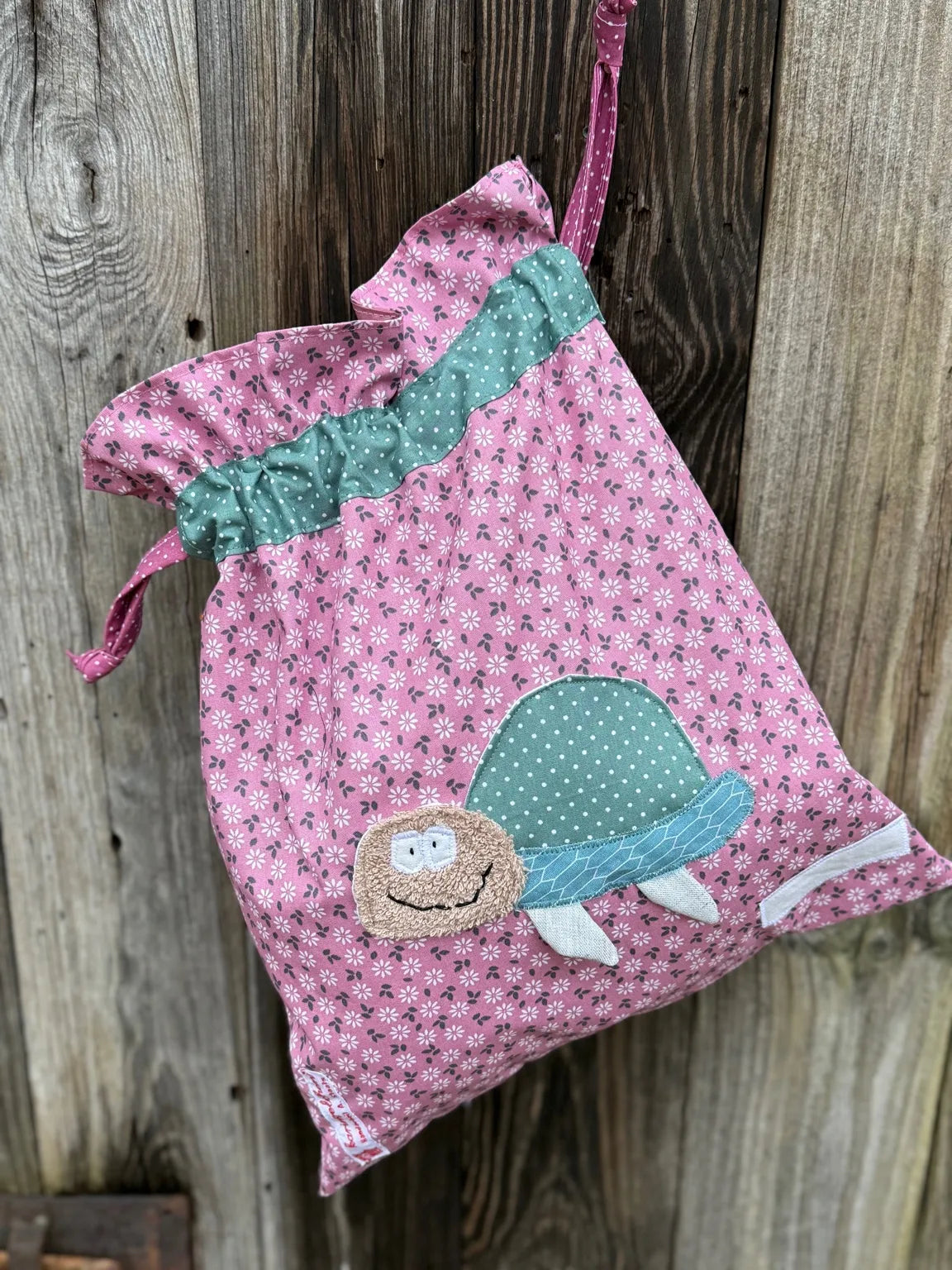Altrosa Wetbag mit weißen Blümchen, verziert mit einer bunten Stoffapplikation einer Schildkröte .DerBeutel hat einen altmint gepunkteten Tunnelzug und altrosa Kordeln, aufgenommen vor rustikalem
Holzhintergrund