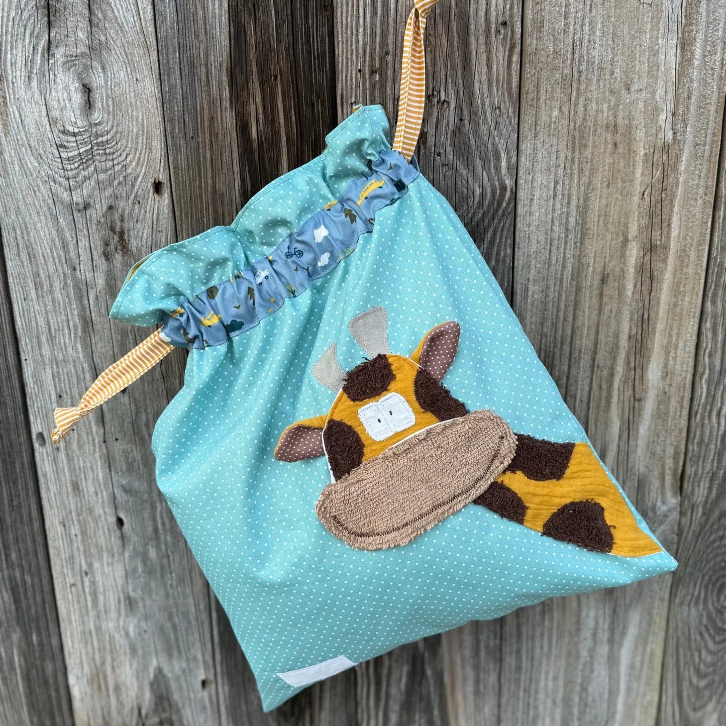 Mintfarbener Wetbag mit weißen Punkten, verziert mit einer bunten Stoffapplikation einer Giraffe .DerBeutel hat einen blauen Tunnelzug und senfgelbeKordeln, aufgenommen vor rustikalem Holzhintergrund.