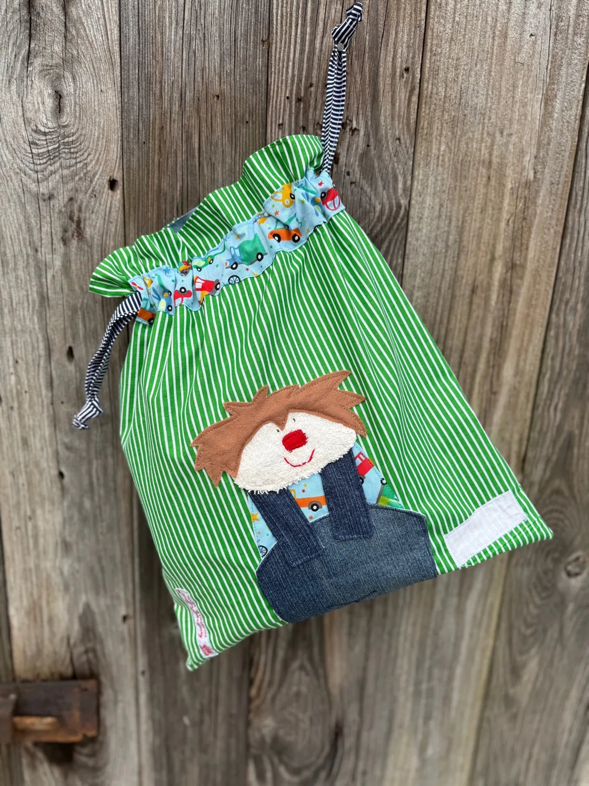 Grüner Wetbag mit weißen Streifen , verziert mit einer bunten Stoffapplikation einen Jungen .DerBeutel hat einen Tunnelzug mit Autoprint und blau gestreiften Kordeln, aufgenommen vor rustikalem Holzhintergrund.