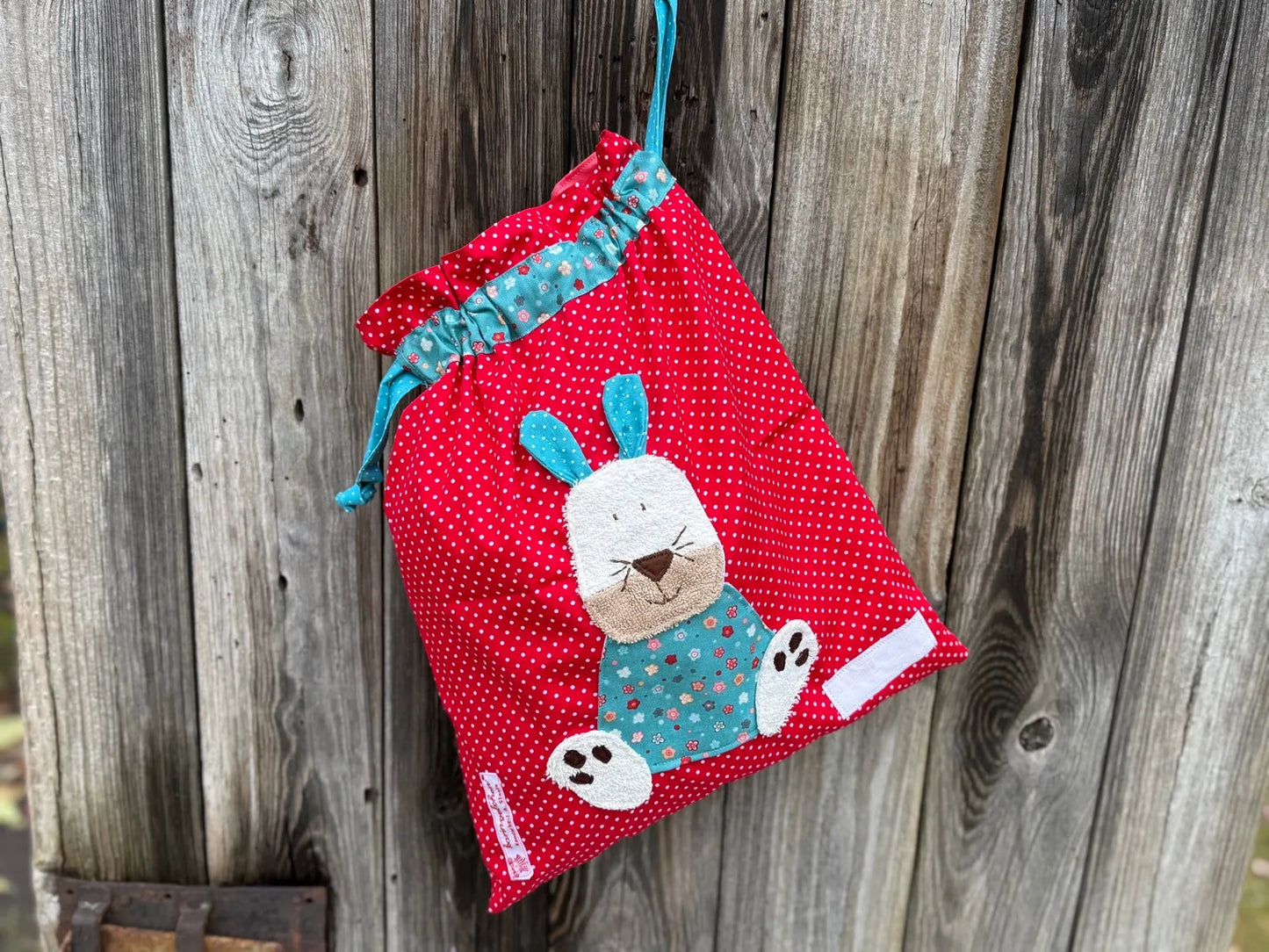 Roter Wetbag mit weißen Punkten, verziert mit einer bunten Stoffapplikation einem Hasen .DerBeutel hat einen floralen Tunnelzug und türkise Kordeln, aufgenommen vor rustikalem Holzhintergrund.
