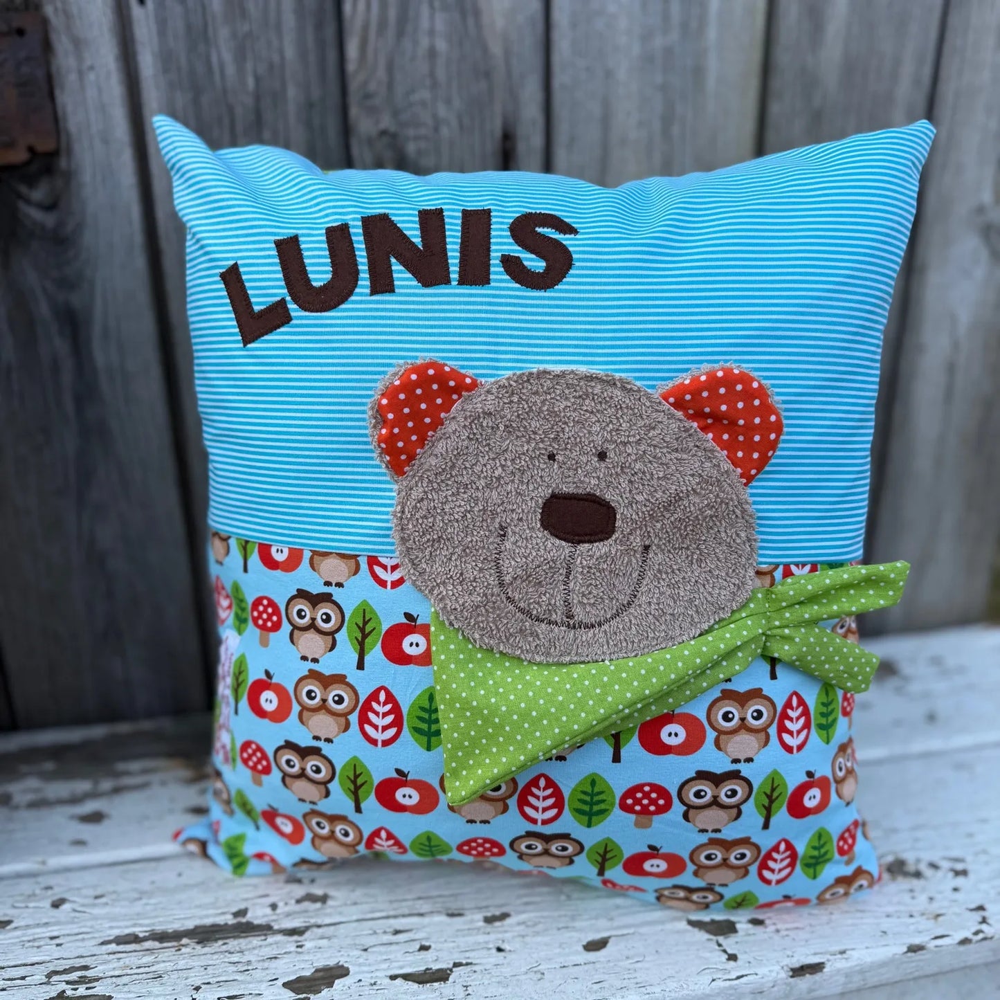 Handgenähtes Kinderkissen personalisiert, gestaltet Türkis mit weißen Streifen im oberen Teil, hellblau mit Print aus Eulen,Blättern und Pilzen im unteren Teil. Auf dem Kissen ist eine Bär-Applikation in hellbraunem Frottee.Fotografiert vor einem rustikalen Holzhintergrund