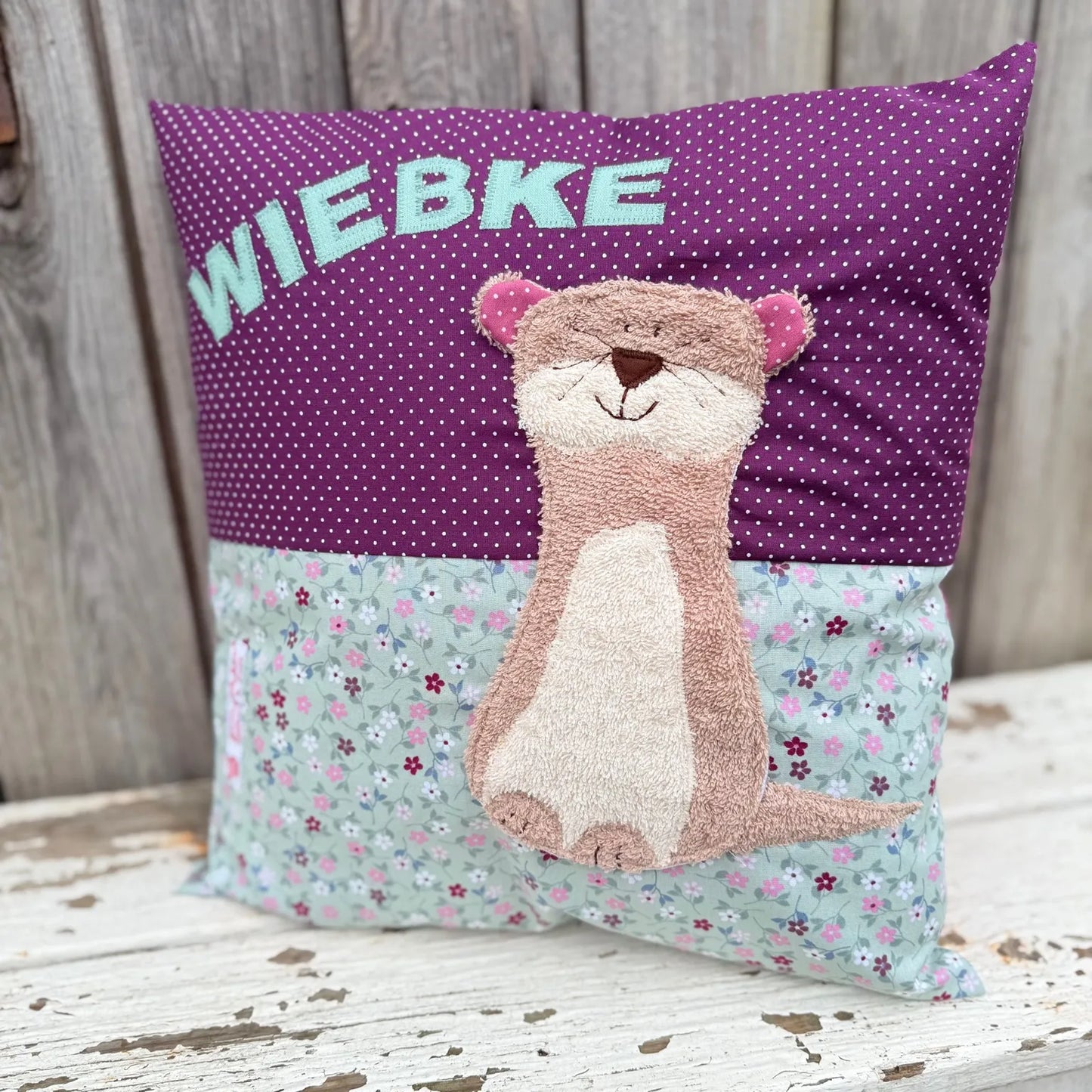 Handgenähtes Kinderkissen personalisiert, gestaltet dunkellila mit weißen Punkten im oberen Teil, altmint mit bunten Blümchen im unteren Teil. Auf dem Kissen ist eine Otter-Applikation in hellbraunem Frottee.Fotografiert vor einem rustikalen Holzhintergrund