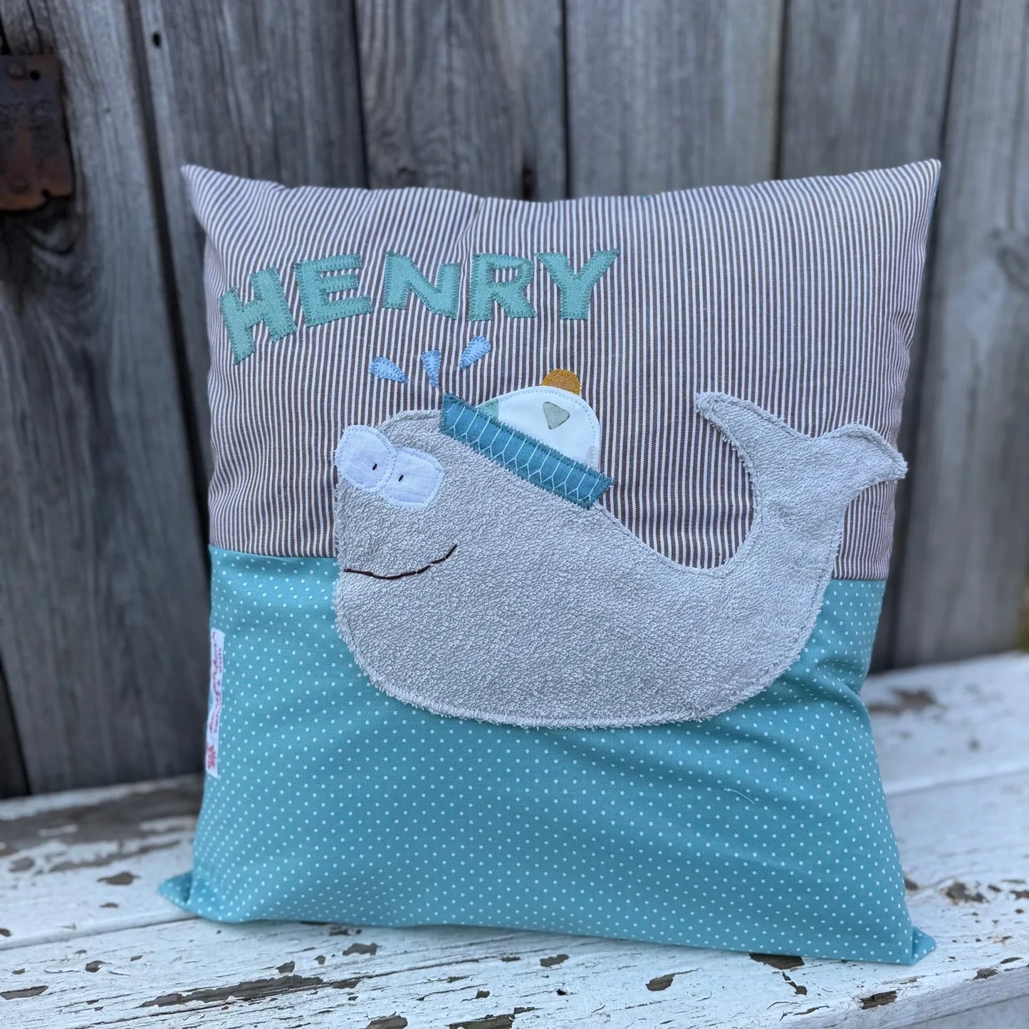Handgenähtes Kinderkissen personalisiert, gestaltet braun- weiß gestreift im oberen Teil, mint mit weißen Punkten im unteren Teil. Auf dem Kissen ist eine Wal-Applikation in grauem Frottee.Fotografiert vor einem rustikalen Holzhintergrund.