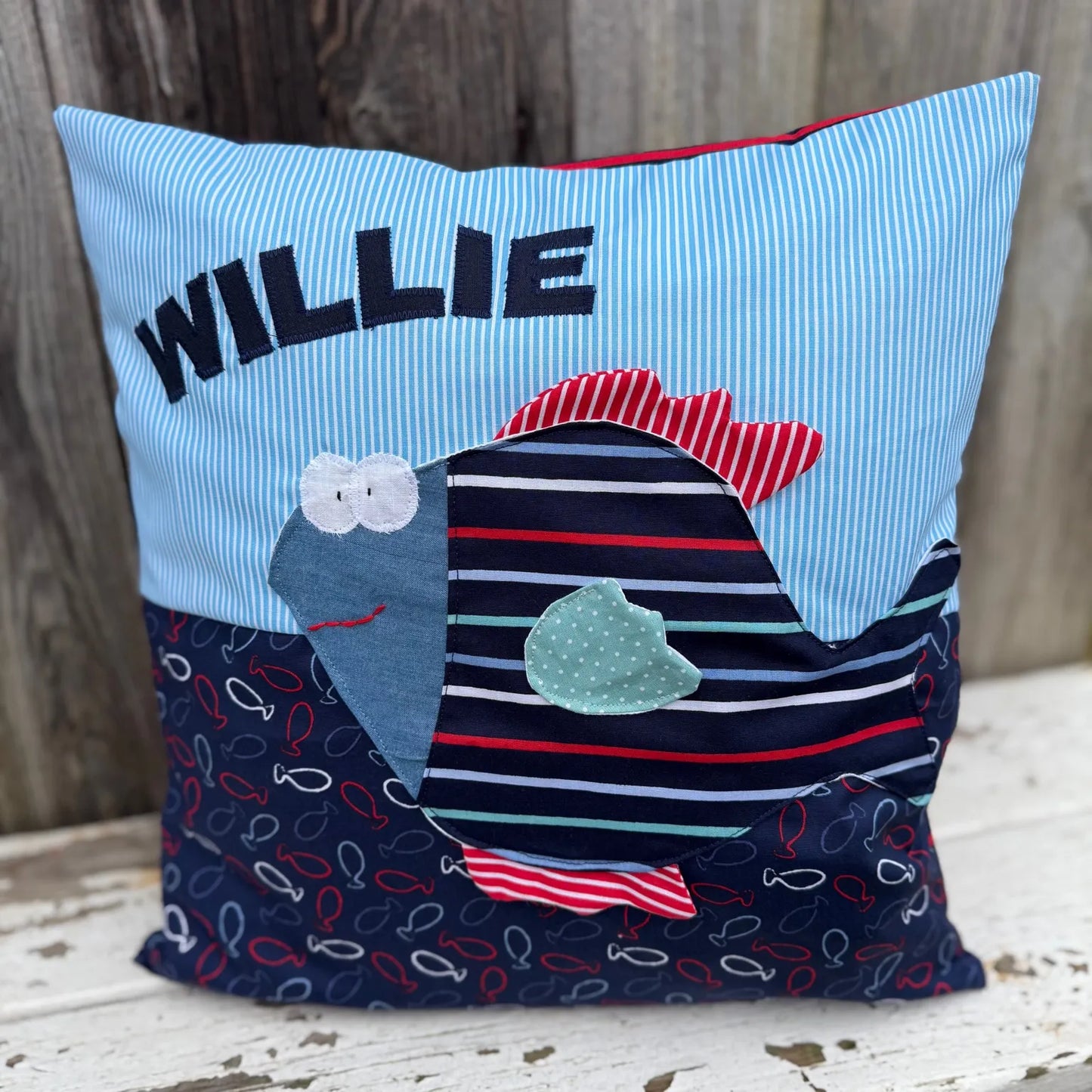 Handgenähtes Kinderkissen personalisiert, gestaltet hellblau mit weißen Streifen im oberen Teil, dunkelblau mit Fischprint im unteren Teil. Auf dem Kissen ist eine Fisch-Applikation im Streifendesign.Fotografiert vor einem rustikalen Holzhintergrund
