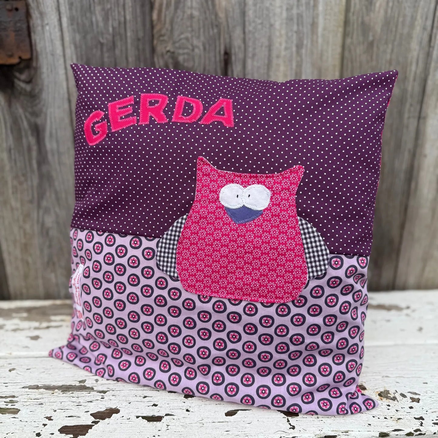 Handgenähtes Kinderkissen personalisiert, gestaltet dunkellila mit weißen Punkten im oberen Teil, lila
mit pinken Blumen im unteren Teil. Auf dem Kissen ist eine Eulen-Applikation in leuchtendem Pink.Fotografiert vor einem rustikalen Holzhintergrund