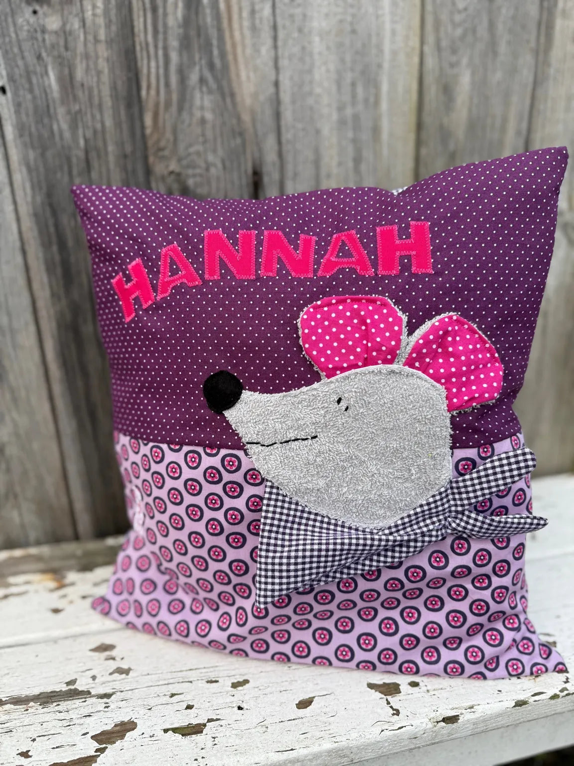 Handgenähtes Kinderkissen personalisiert, gestaltet dunkellila mit weißen Punkten im oberen Teil, lila mit pinken Blumen im unteren Teil. Auf dem Kissen ist einer Maus-Applikation in grauem Frottee. Fotografiert vor einem rustikalen Holzhintergrund