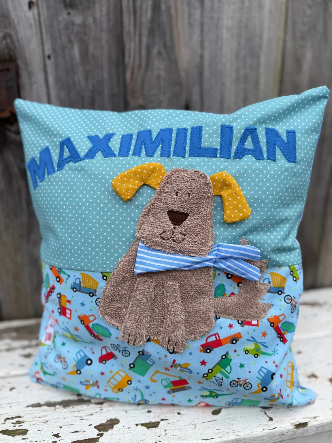Handgenähtes Kinderkissen personalisiert, gestaltet mint mit weißen Punkten im oberen Teil, hellblau mit bunten Fahrzeugen im unteren Teil. Auf dem Kissen ist eine Hunde-Applikation in hellbraunem Frottee . Fotografiert vor einem rustikalen Holzhintergrund