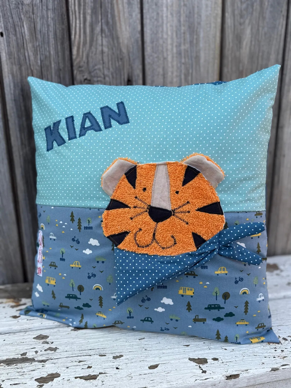 Handgenähtes Kinderkissen personalisiert, gestaltet mint mit weißen Punkten im oberen Teil, blauen mit
Autoprint im unteren Teil. Auf dem Kissen ist eine Tiger-Applikation in leuchtendem Orange. Fotografiert vor einem rustikalen Holzhintergrund