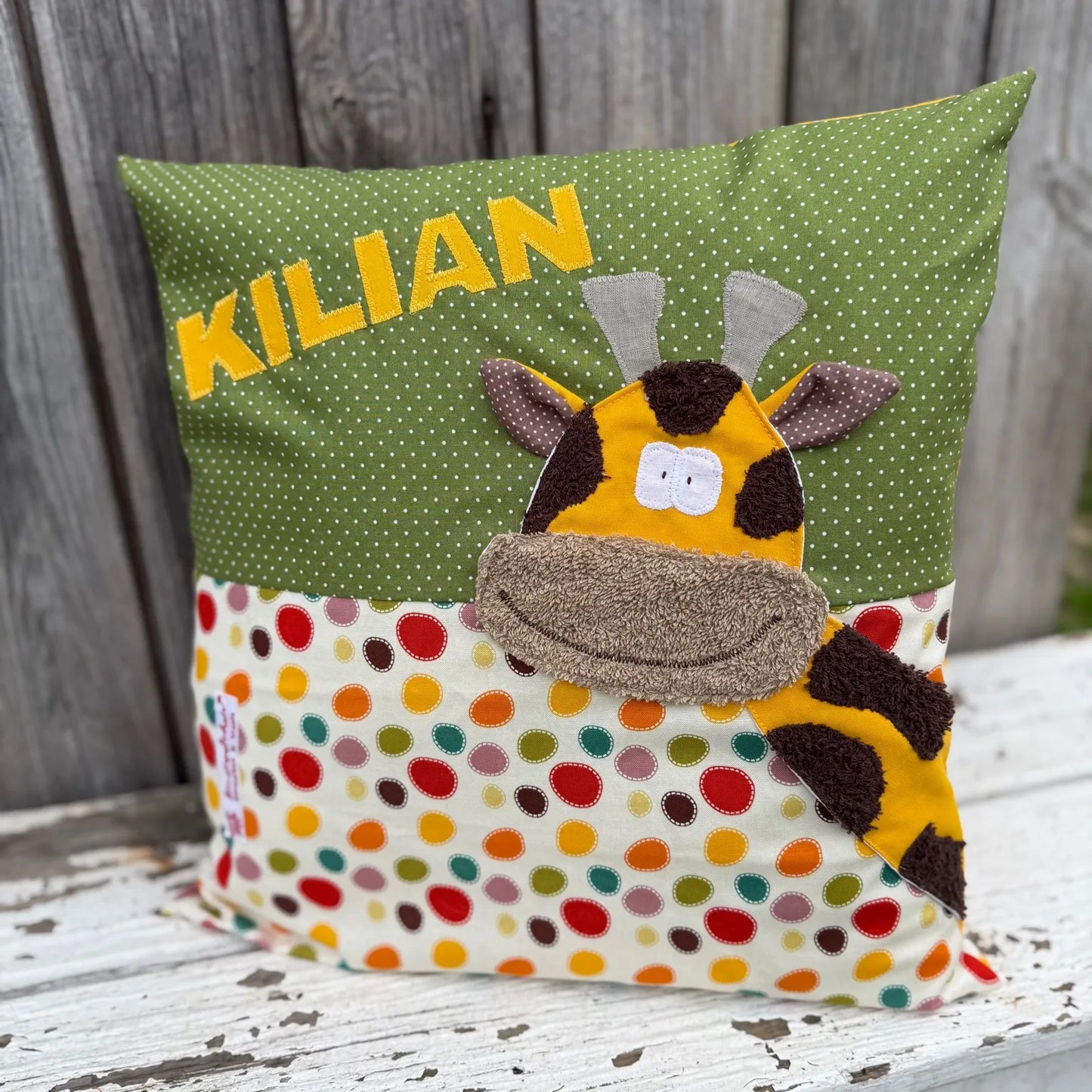 Handgenähtes Kinderkissen personalisiert, gestaltet grün mit weißen Punkten im oberen Teil, cremefarben mit bunten Tupfen im unteren Teil. Auf dem Kissen ist eine Giraffen-Applikation in leuchtendem Gelb. Fotografiert vor einem rustikalen Holzhintergrund