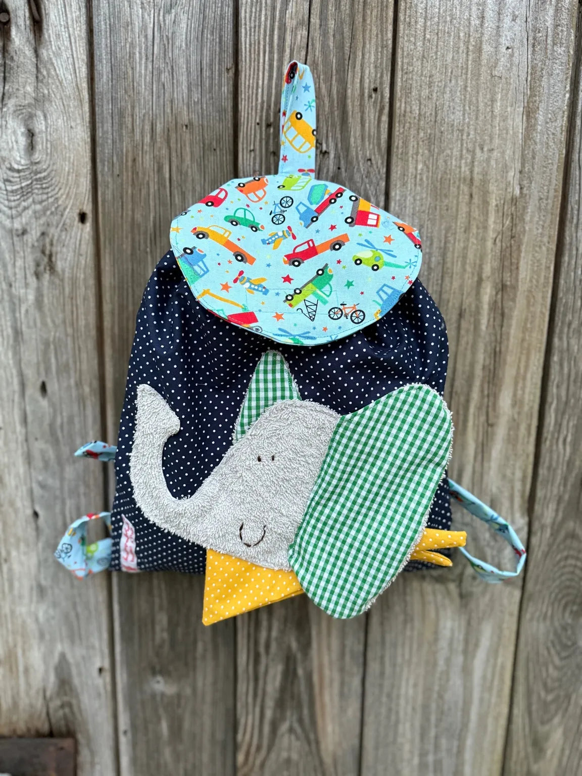 Handgemachter Kinderrucksack
ist dunkelblau mit weißen Punkten , hat eine hellblaue Klappe mit bunten
Fahrzeugeprint und eine aufgenähte Applikation eines
grauen Elefanten.
