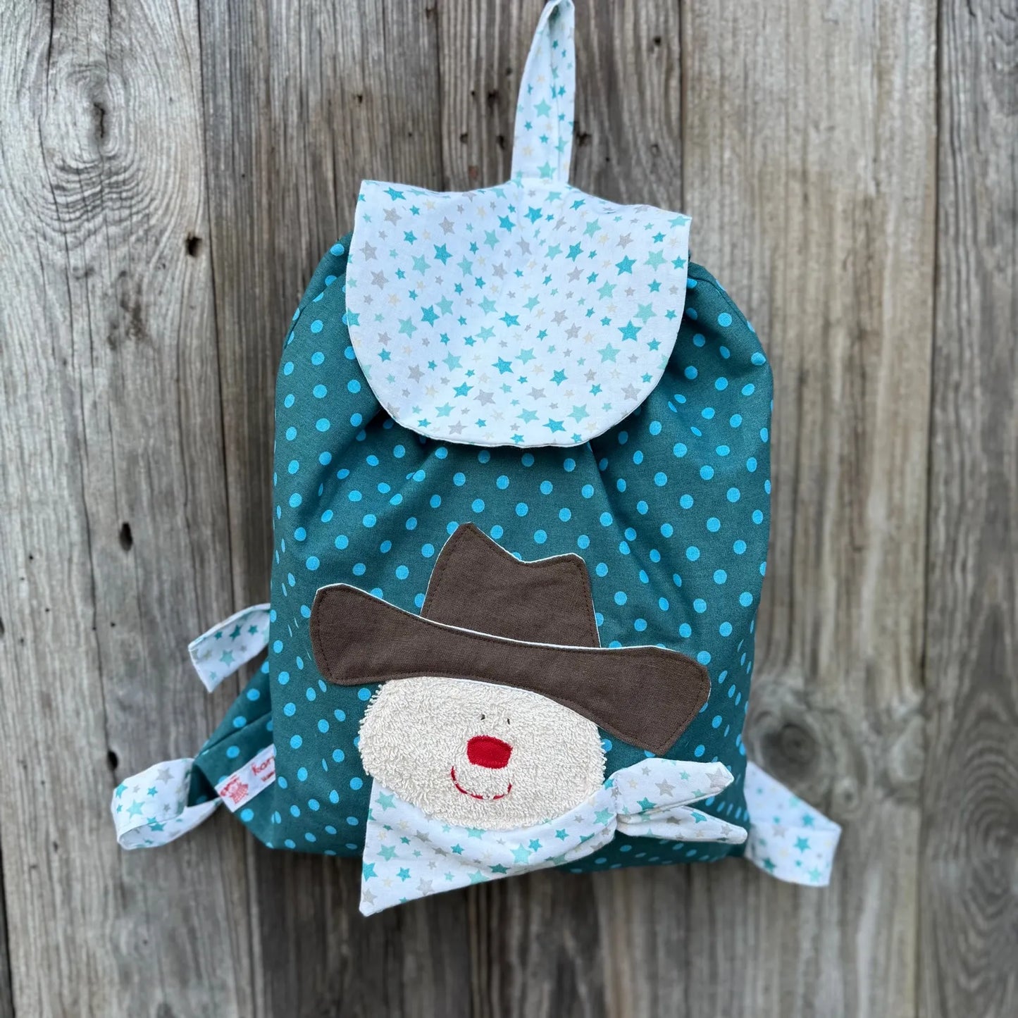 Handgemachter Kinderrucksack ist petrol
mit Punkten , hat eine weiße Klappe mit Sternen und eine aufgenähte Applikation eines Cowboys.