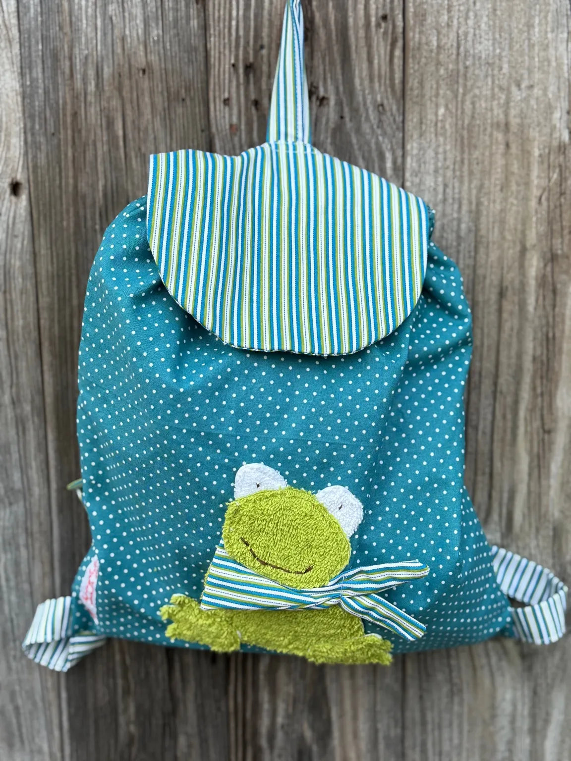 Handgemachter Kinderrucksack
ist petrolmit weißen Punkten , hat eine grün petrol gestreifte Klappe
und eine aufgenähte Applikation eines
grünen Froschs.