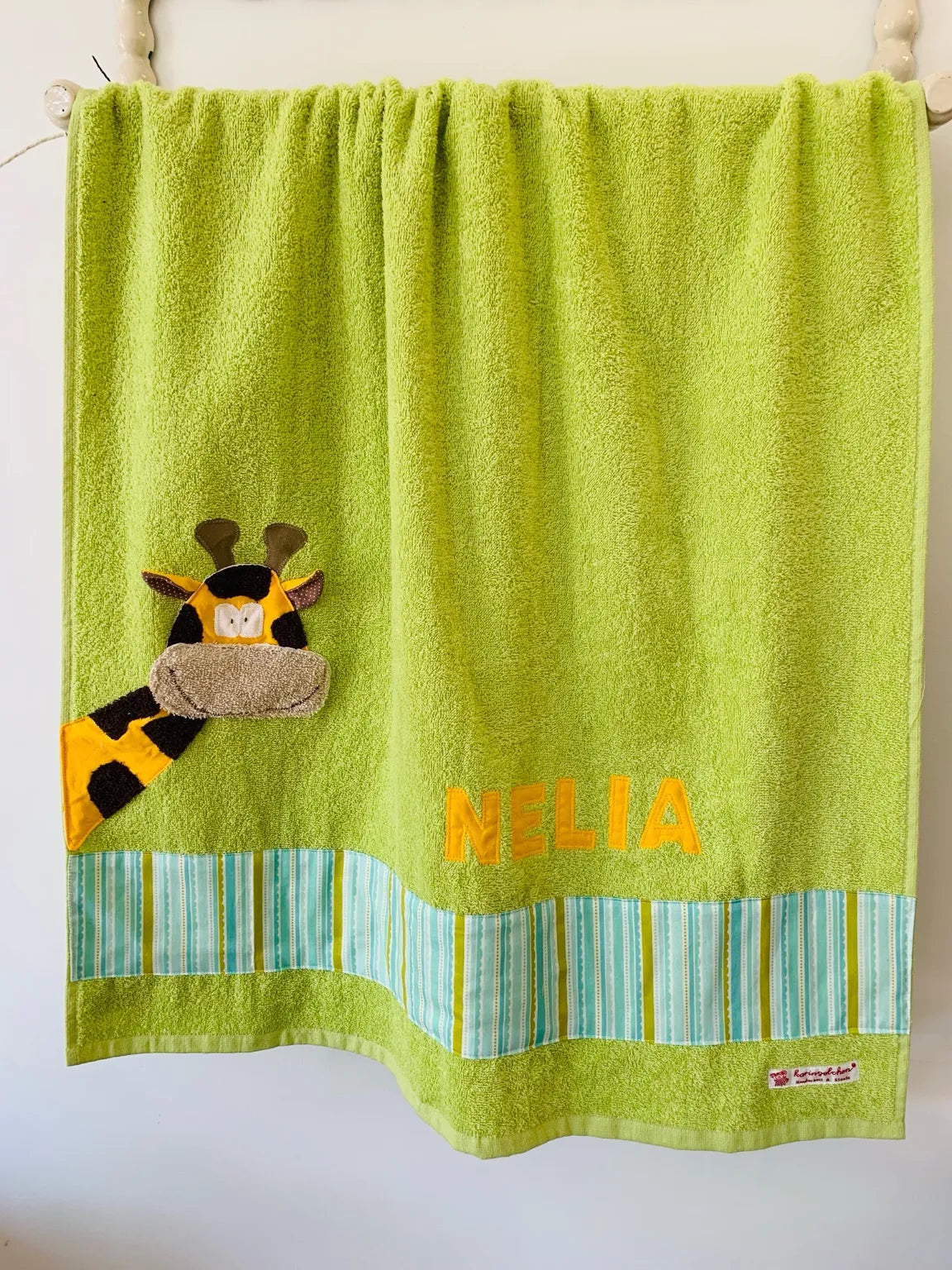Handgemachtes hellgrünes Duschtuch mit applizierter Giraffe in der linken unteren Ecke, personalisiert mit Namen in gelben Buchstaben. Unten am Handtuch ist ein Streifen in grün blau gestreift aufgenäht.