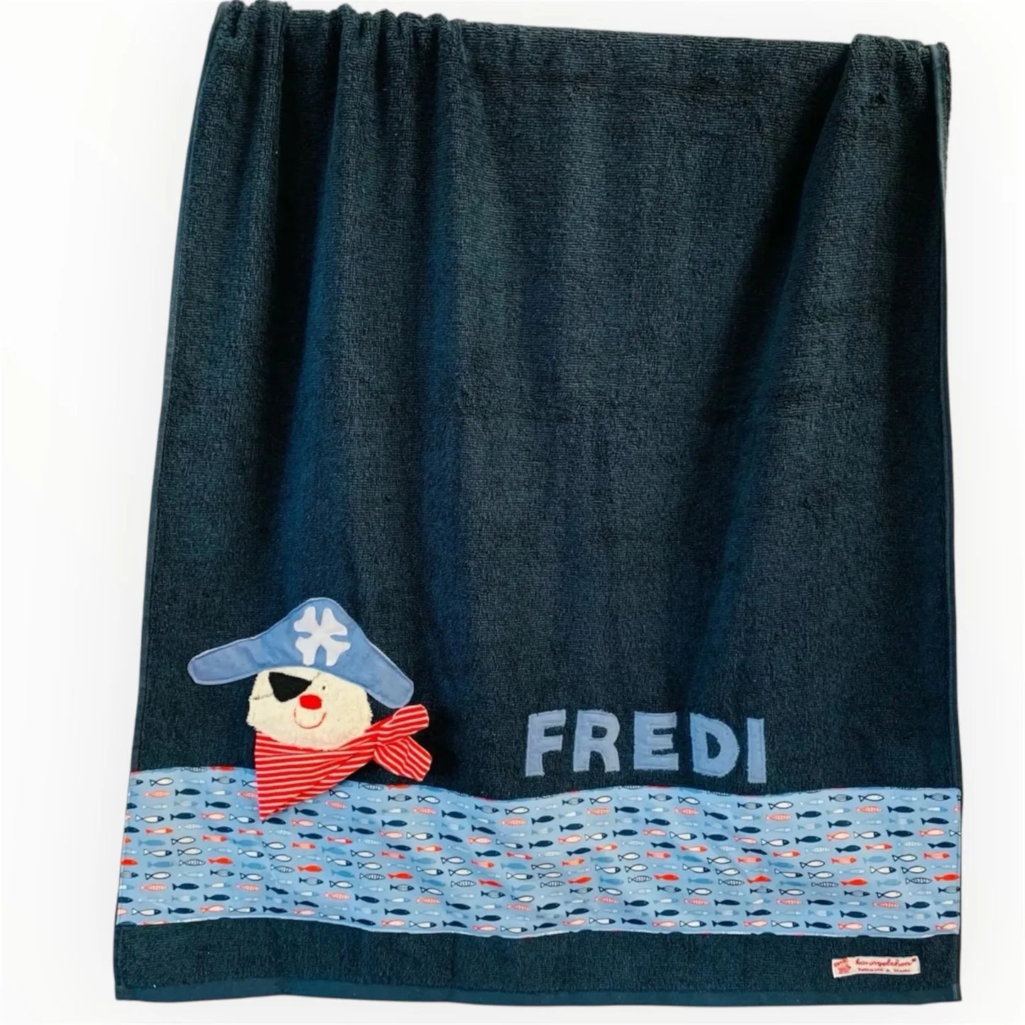 Handgemachtes dunkelblaues Duschtuch mit appliziertem Pirat in der linken unteren Ecke, personalisiert mit Namen in hellblauen Buchstaben. Unten am Handtuch ist ein Streifen in hellblauen mit Sardinen aufgenäht.