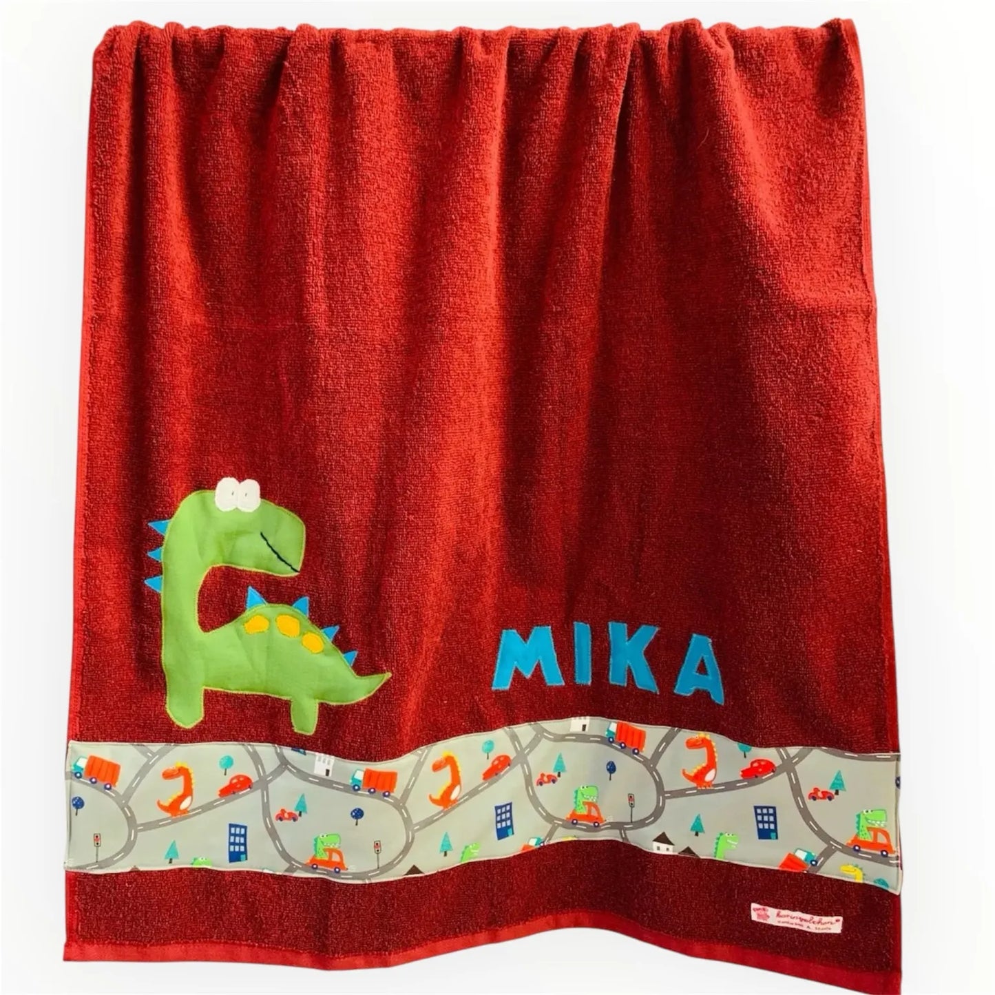 Handgemachtes bordeauxrotesDuschtuch mit appliziertem grünen Dino in der linken unteren Ecke, personalisiert mit Namen in hellblauen Buchstaben. Unten am Handtuch ist ein Streifen in grau mit Dinos im Straßenverkehr aufgenäht.