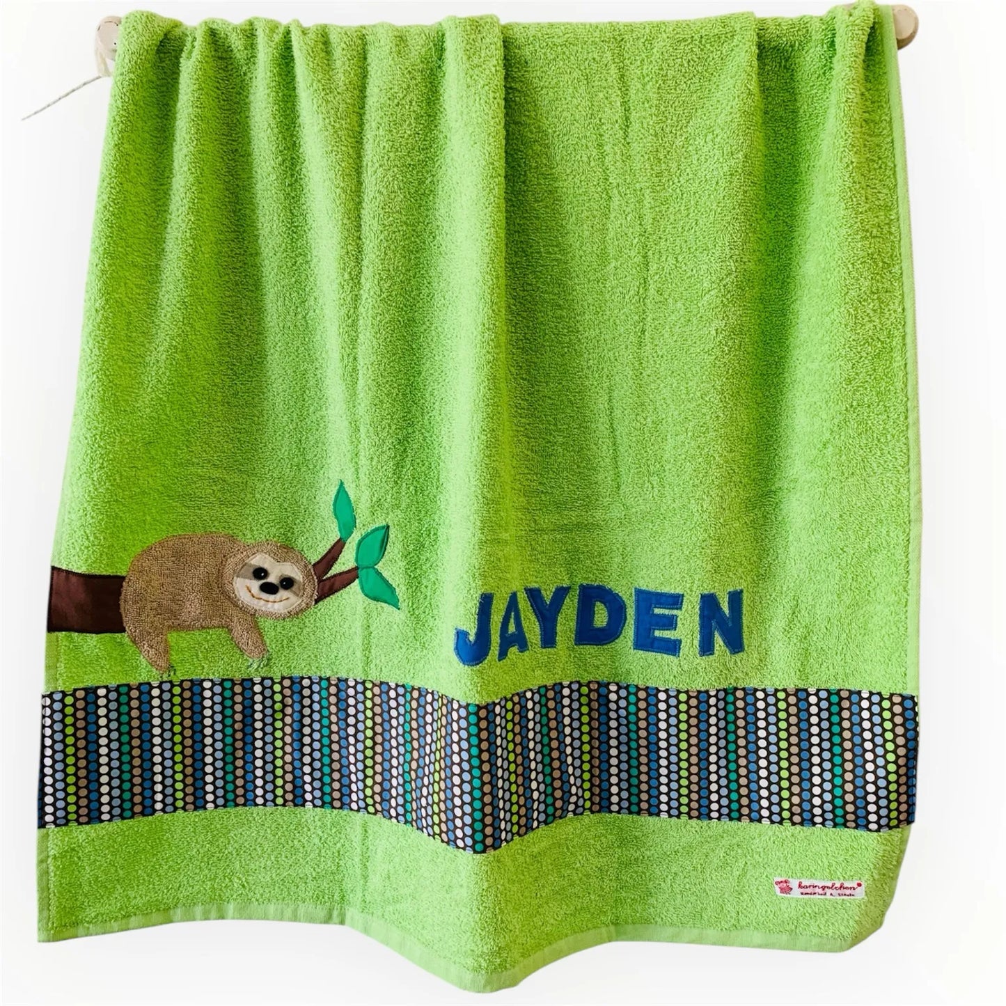 Handgemachtes hellgrünes Duschtuch mit appliziertem Faultier in der linken unteren Ecke, personalisiert mit Namen in gelben Buchstaben. Unten am Handtuch ist ein Streifen in braun grün blau gepunktet aufgenäht.