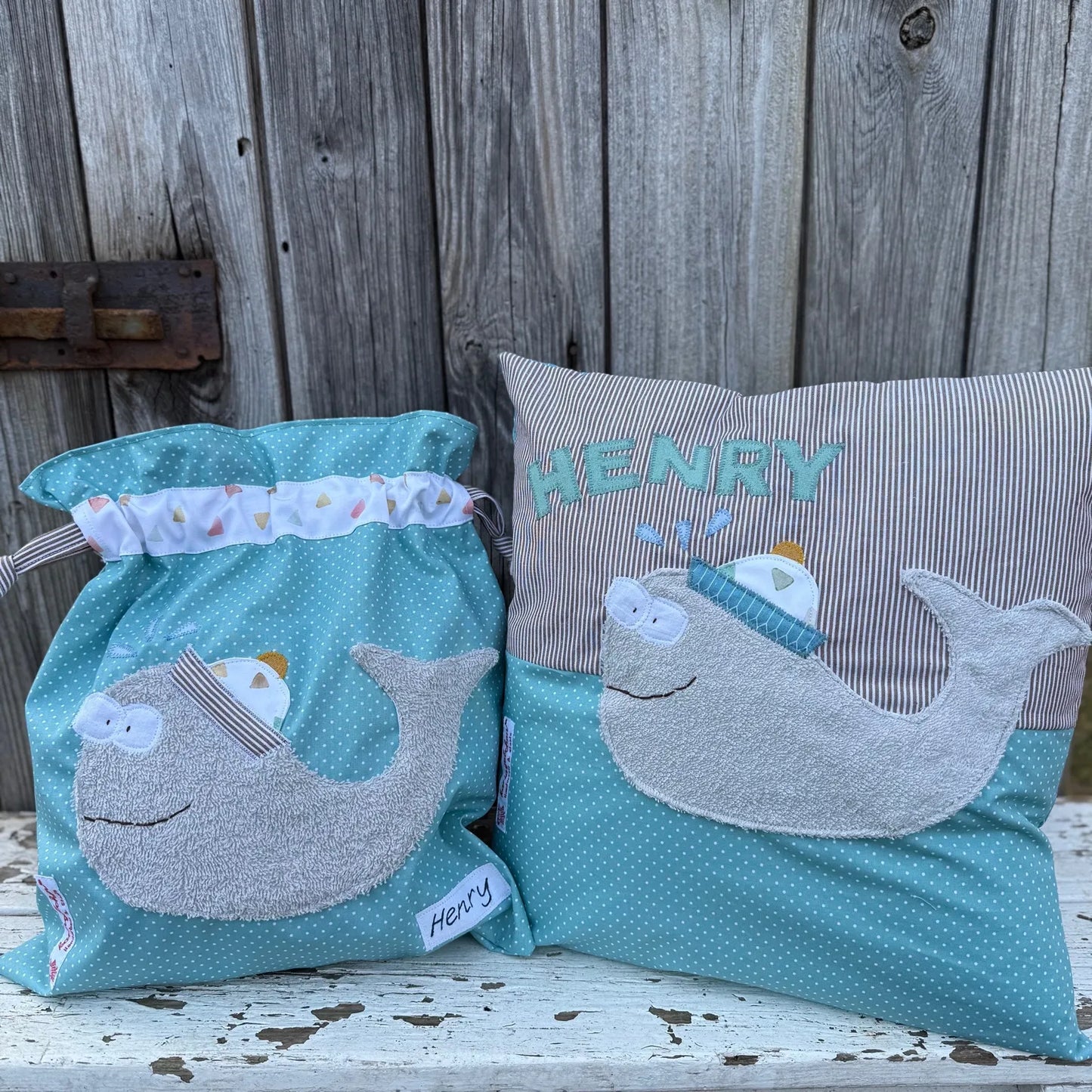 Handgefertigtes Set bestehend aus einer Wetbag und einem personalisierten Kissen. Beide Artikel sind aus türkisfarbenem Stoff mit weißen Punkten sowie braun-weiß gestreiftem Stoff gefertigt. Auf beiden ist eine freundliche Wal-Applikation mit Kapitänsmütze und Brille zu sehen. Das Kissen trägt den Namen „HENRY“ in türkisfarbenen Buchstaben. Die Wetbag hat einen Tunnelzug mit maritim gemustertem Stoff. Die Artikel stehen auf einer weiß gestrichenen, verwitterten Holzbank vor einer rustikalen Holzwand.