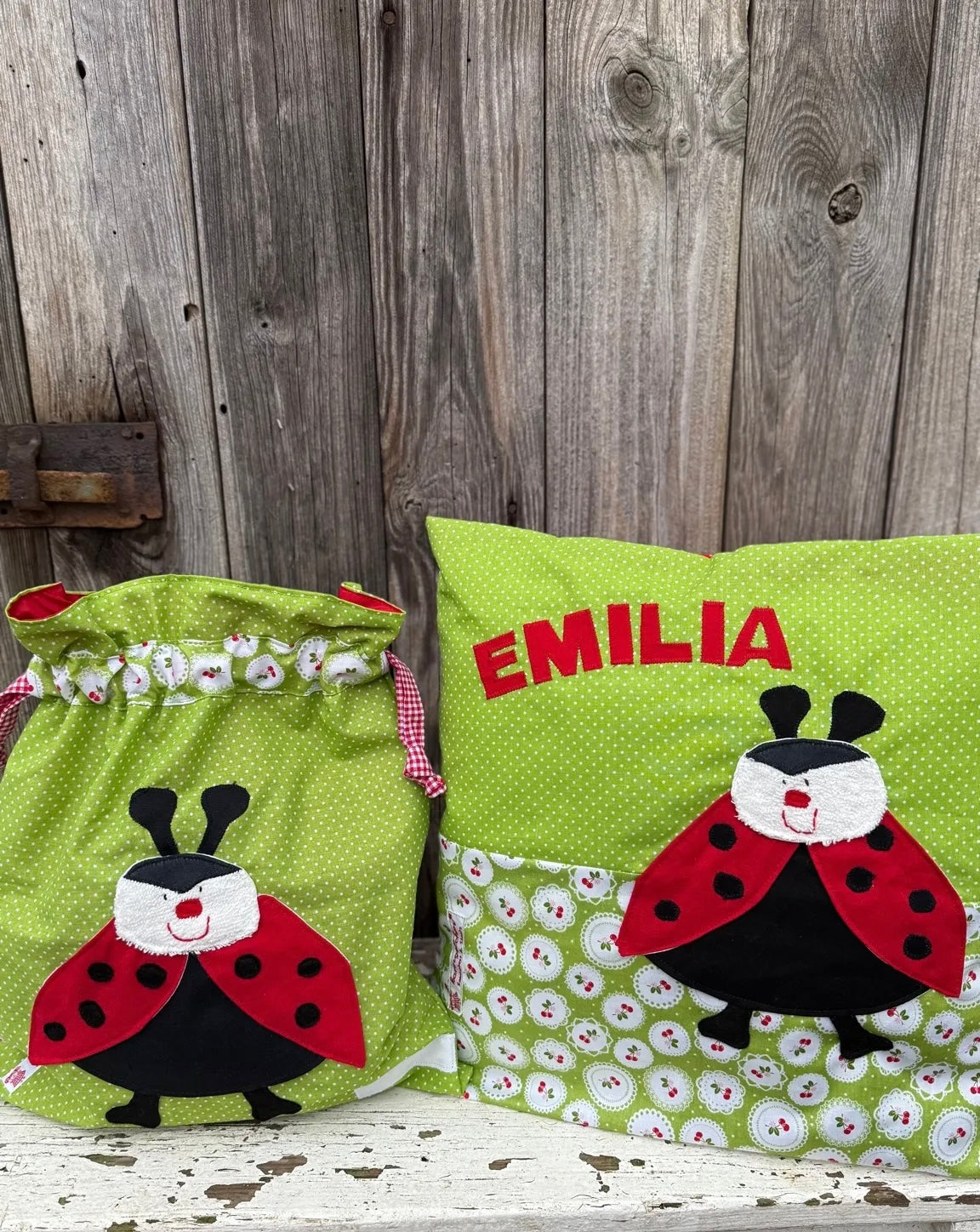 Handgefertigtes Set aus Wetbag und personalisiertem Kissen in fröhlichem Design. Beide Teile bestehen aus grünem Stoff mit weißen Punkten und sind mit einer Marienkäfer-Applikation versehen. Die Marienkäfer haben einen schwarzen Körper, rote Flügel mit schwarzen Punkten und ein weißes Gesicht mit rotem Mund. Das Kissen ist zusätzlich mit dem Namen „EMILIA“ in roten Buchstaben personalisiert. Die Artikel stehen auf einer weiß gestrichenen, verwitterten Holzbank vor einer rustikalen Holzwand.