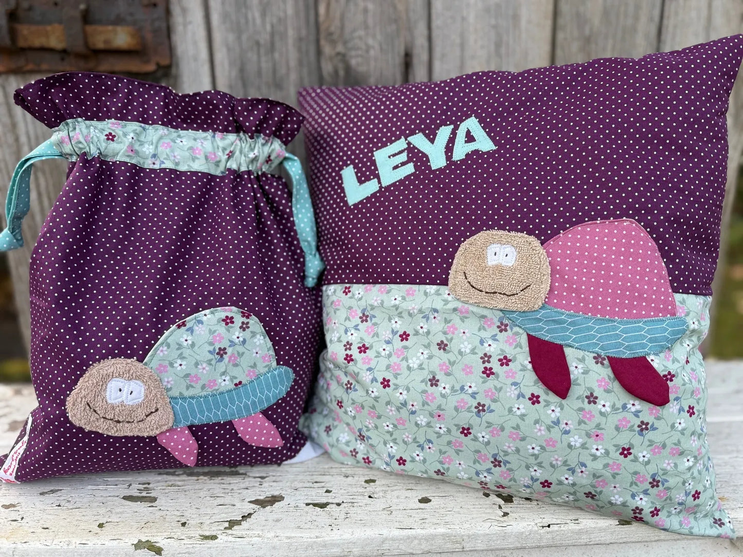 Handgefertigtes Set aus einer Wetbag und einem personalisierten Kissen. Beide Stücke bestehen aus weinrotem Stoff mit weißen Punkten und einem floralen Stoff in Mintgrün. Auf beiden ist eine applizierte Schildkröte mit fröhlichem Gesicht und buntem Panzer zu sehen. Das Kissen ist zusätzlich mit dem Namen „LEYA“ in hellblauen Buchstaben personalisiert. Die Produkte stehen auf einer weißen Holzbank vor einer rustikalen Holzwand.