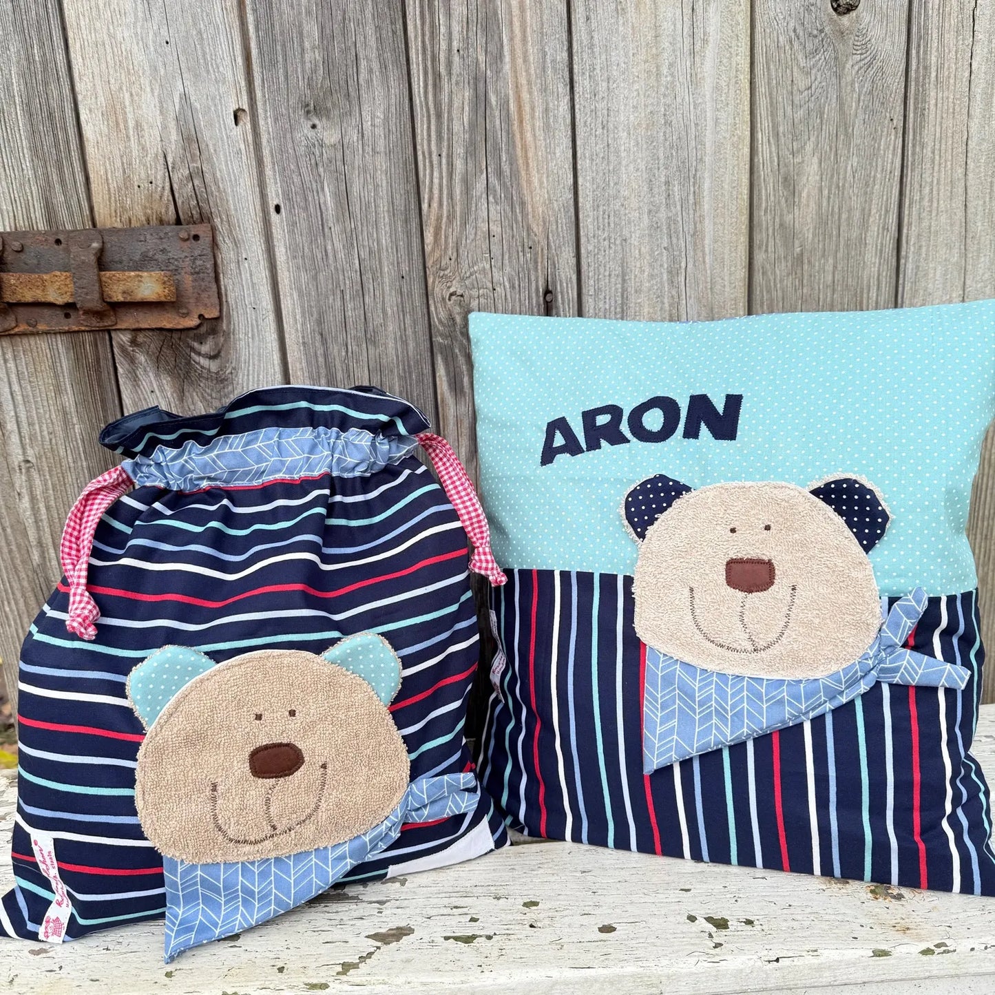 Handgemachtes Set aus einer Wetbag und einem personalisierten Kissen. Beide Artikel zeigen eine freundliche Bären-Applikation mit beigem Gesicht, bunten Ohren und blauem Halstuch. Die Wetbag ist aus dunkelblauem Stoff mit weißen und roten Streifen sowie rosa Kordelzug gefertigt. Das Kissen besteht aus hellblauem Punktestoff oben und gestreiftem Stoff unten. Der Name „ARON“ ist in blauen Buchstaben auf das Kissen appliziert. Die Artikel stehen auf einer weiß gestrichenen, verwitterten Holzbank vor ein