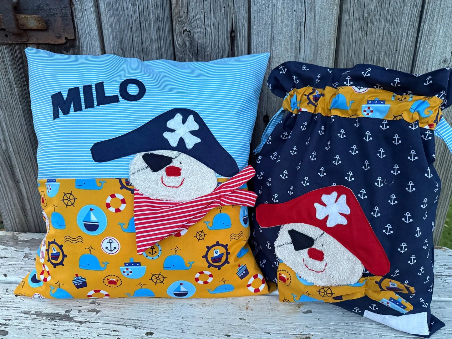 Handgefertigtes Set bestehend aus einem personalisierten Kissen und einer Wetbag im maritimen Piraten-Design. Das Kissen hat einen hellblauen, gestreiften oberen Teil und einen unteren Teil mit gelbem Stoff, bedruckt mit Schiffen, Ankern und Rettungsringen. Beide Teile zeigen eine fröhliche Piraten-Applikation mit Augenklappe, rotem Kopftuch und Piratenhut. Auf dem Kissen ist der Name „MILO“ in dunkelblauen Buchstaben aufgestickt. Die Wetbag ist aus dunkelblauem Ankerstoff gefertigt, mit gelbem Motivst