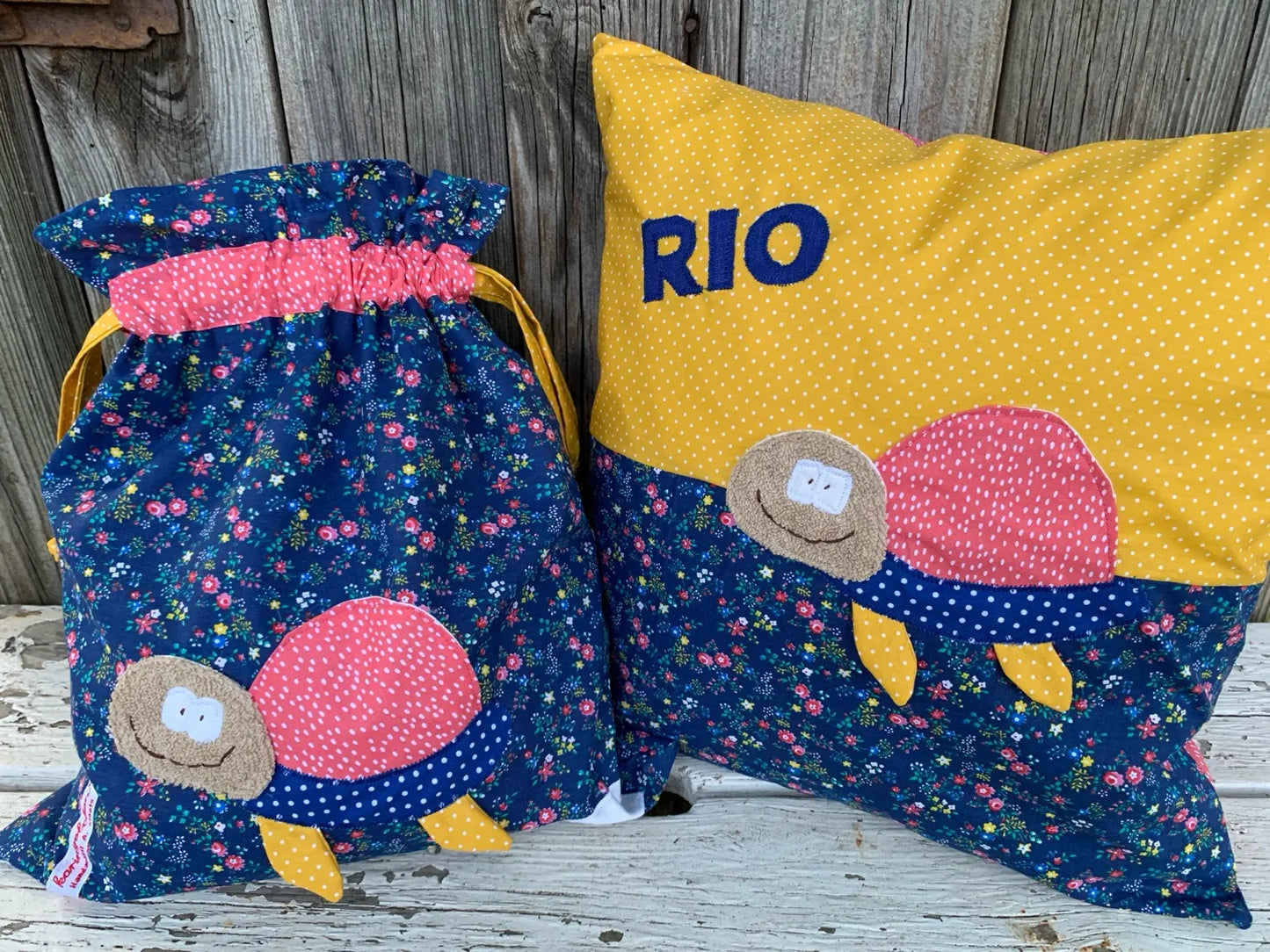 Handgefertigtes Set bestehend aus einer Wetbag und einem personalisierten Kissen. Beide Artikel sind aus dunkelblauem Stoff mit buntem Blumenmuster sowie gelbem und pink gepunktetem Stoff gearbeitet. Appliziert ist eine fröhliche Schildkröte mit beigem Kopf, pinkem Panzer, blauem Körper mit weißen Punkten und gelben Beinen. Das Kissen ist mit dem Namen „RIO“ in blauen Buchstaben personalisiert. Die Produkte stehen auf einer weißen Holzbank vor einer rustikalen Holzwand.