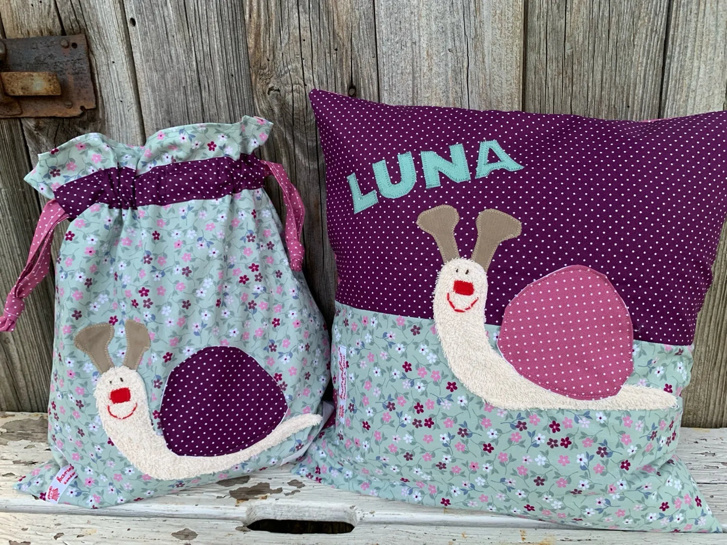 Handgemachtes Set bestehend aus einer Wetbag und einem personalisierten Kissen. Beide sind aus mintgrünem Stoff mit Blumenmuster und weinrotem Stoff mit weißen Punkten gefertigt. Sowohl auf der Wetbag als auch auf dem Kissen ist eine applizierte Schnecke mit fröhlichem Gesicht und gepunktetem Haus aufgenäht. Das Kissen trägt zusätzlich den Namen „LUNA“ in türkisfarbenen Buchstaben. Die Artikel stehen auf einer weiß gestrichenen, rustikalen Holzbank vor einer verwitterten Holzwand.