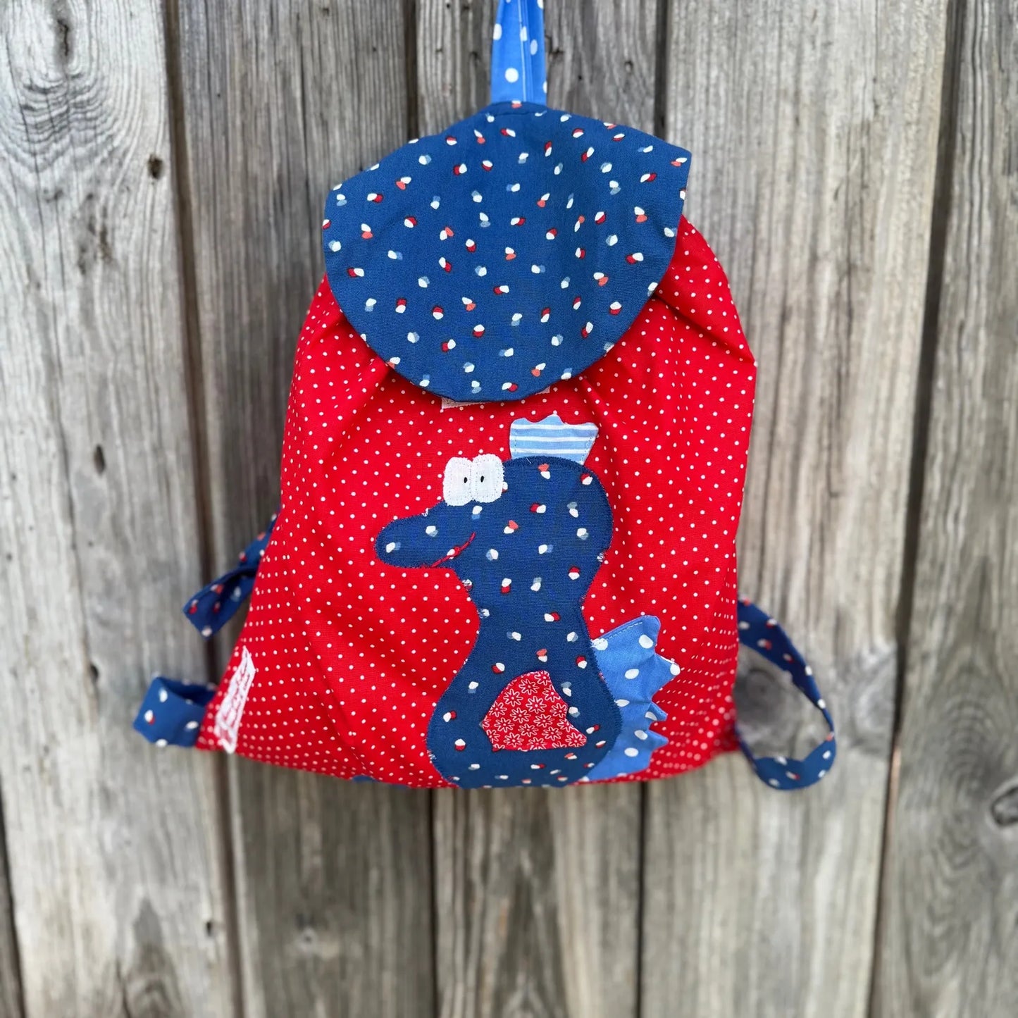 Handgemachter Kinderrucksack ist rot mit
mit weißen Punkten , hat eine Royalblauer Klappe mit bunten Punkten und eine aufgenähte Applikation eines blauen Seepferdchens.