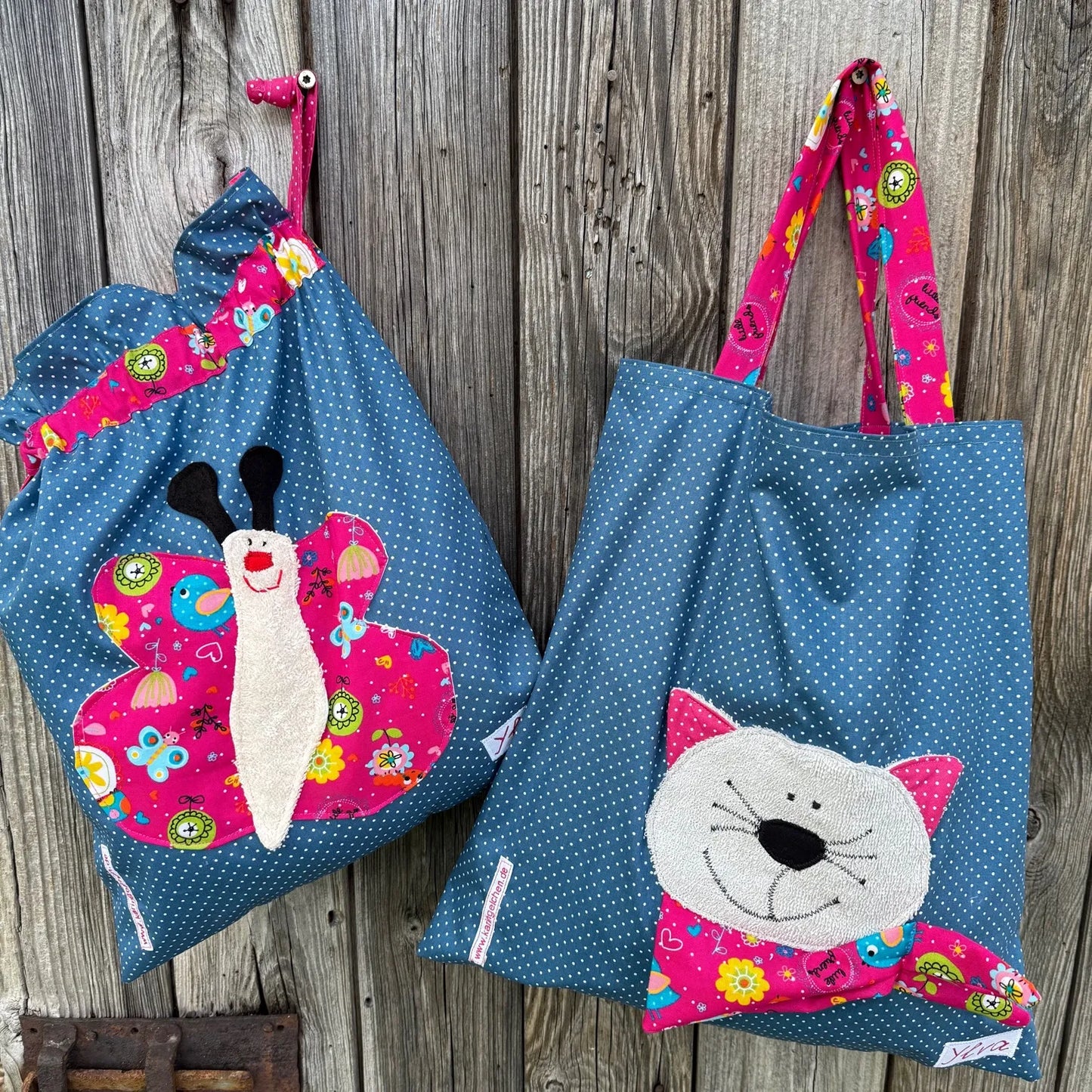 “KitaSet Handmade bestehend aus einem blauen Garderobenbeutel mit pink gemusterter Schmetterlings-Applikation aus Frottee im 3D-Effekt und einer passenden Stofftasche mit Katzenmotiv aus Frottee und bunten Stoffakzenten.”