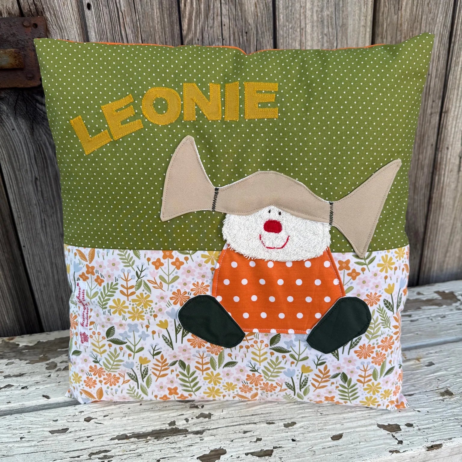 „Handgenähtes Kuschelkissen für Kinder mit grünem Punktestoff, bunter Blumenwiese, appliziertem Mädchen in orangem Punkteshirt und personalisiertem Namen ‚Leonie‘.“