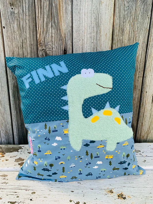 Handgenähtes Kinderkissen personalisiert, gestaltet petrol mit weißen Punkten im oberen Teil, rauchblau mit mit Autoprint im unteren Teil. Auf dem Kissen ist eine Dino-Applikation in mintgrünem Frottee.Fotografiert vor einem rustikalen Holzhintergrund 