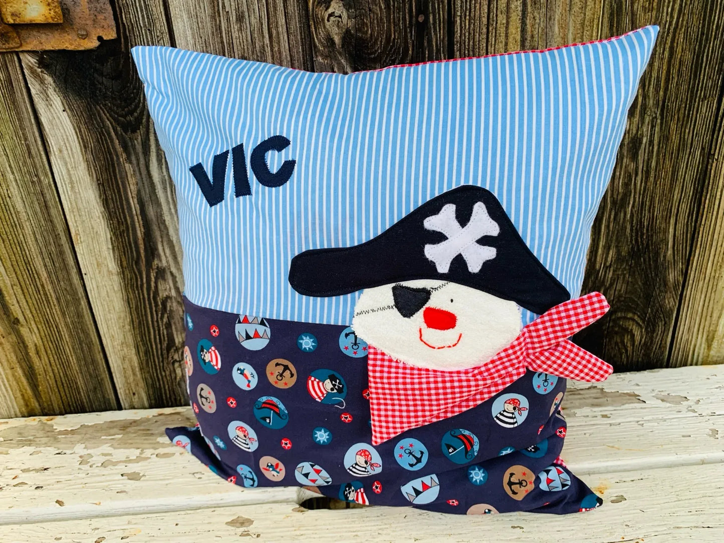 Handgenähtes Kinderkissen personalisiert, gestaltet hellblau mit weißen Streifen im oberen Teil,
 dunkelblau mit Piratenmotiven im unteren Teil. Auf dem Kissen ist eine Piraten-Applikation , fotografiert vor einem rustikalen Holzhintergrund