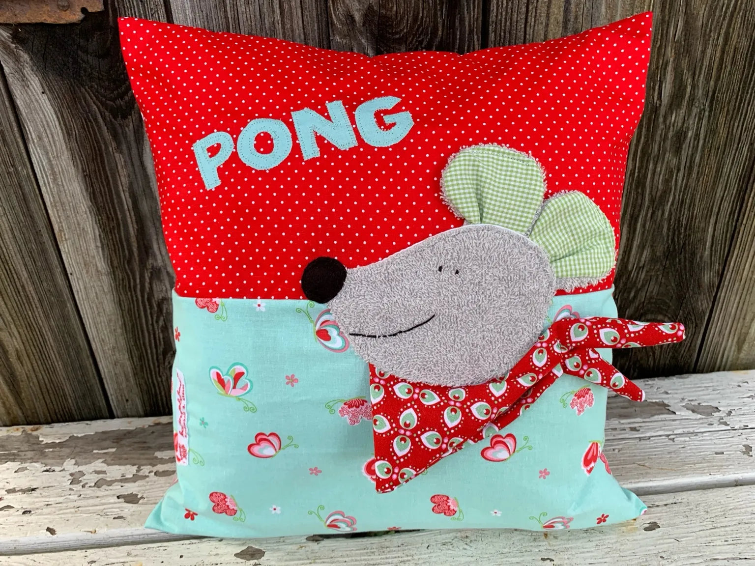 Handgenähtes Kinderkissen personalisiert, gestaltet rot mit weißen Punkten im oberen Teil, Ming mit Schmetterlingprint im unteren Teil. Auf dem Kissen ist eine Maus-Applikation in hellgrauen Frottee.Fotografiert vor einem rustikalen Holzhintergrund 