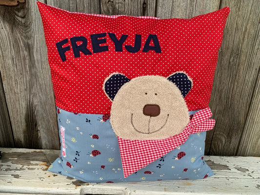 Handgenähtes Kinderkissen personalisiert, gestaltet rot mit weißen Punkten im oberen Teil, rauchblau 
mit Marienkäferprint im unteren Teil. Auf dem Kissen ist eine Bär-Applikation in hellbraunem Frottee.Fotografiert vor einem rustikalen Holzhintergrund 