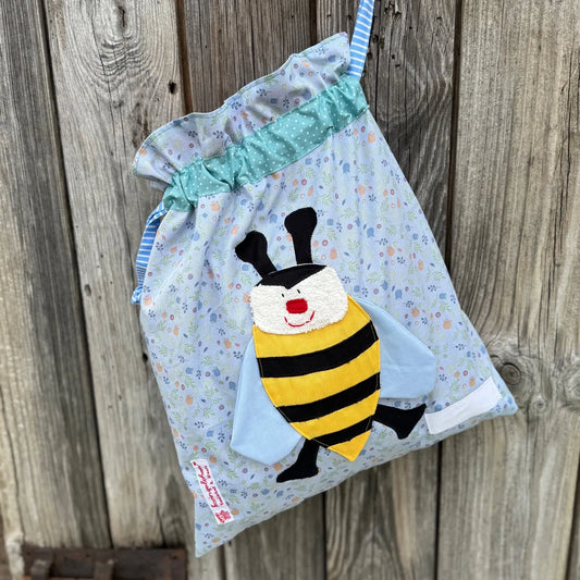 Hellblauer Wetbag mit  pastellfarbenen Blümchen , verziert mit einer bunten Stoffapplikation einer Biene .DerBeutel hat einen mint gepunkteten Tunnelzug und hellblaue Kordeln, aufgenommen vor rustikalem Holzhintergrund. 