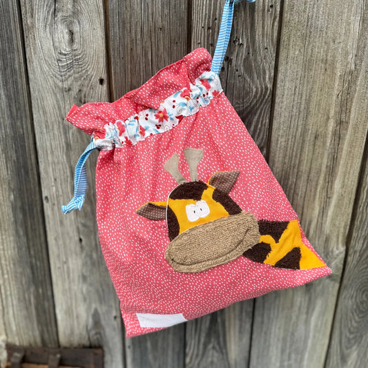 Lachsroter Wetbag mit weißen Punkten, verziert mit einer bunten Stoffapplikation einer Giraffe .DerBeutel hat einen floralen Tunnelzug und hellblaue Kordeln, aufgenommen vor rustikalem Holzhintergrund.