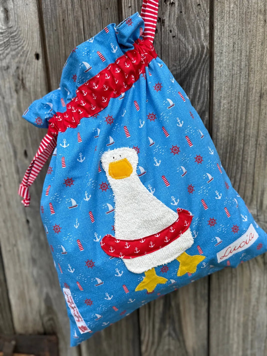 Blauer Wetbag im maritimen Design, verziert mit einer bunten Stoffapplikation einer Ente mit Schwimmreifen .DerBeutel hat einen Tunnelzug aus rotem 
Ankerstoff und rot gestreifte Kordeln, aufgenommen vor rustikalem Holzhintergrund.