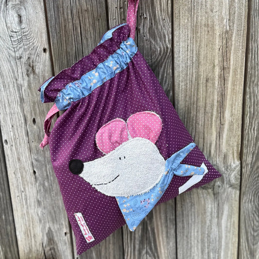 Hellgrüner Wetbag mit weißen Punkten, verziert mit einer bunten Stoffapplikation einer Maus aus grauem Frottee .DerBeutel hat einen floralen Tunnelzug und altrosa Kordeln, aufgenommen vor rustikalem Holzhintergrund.