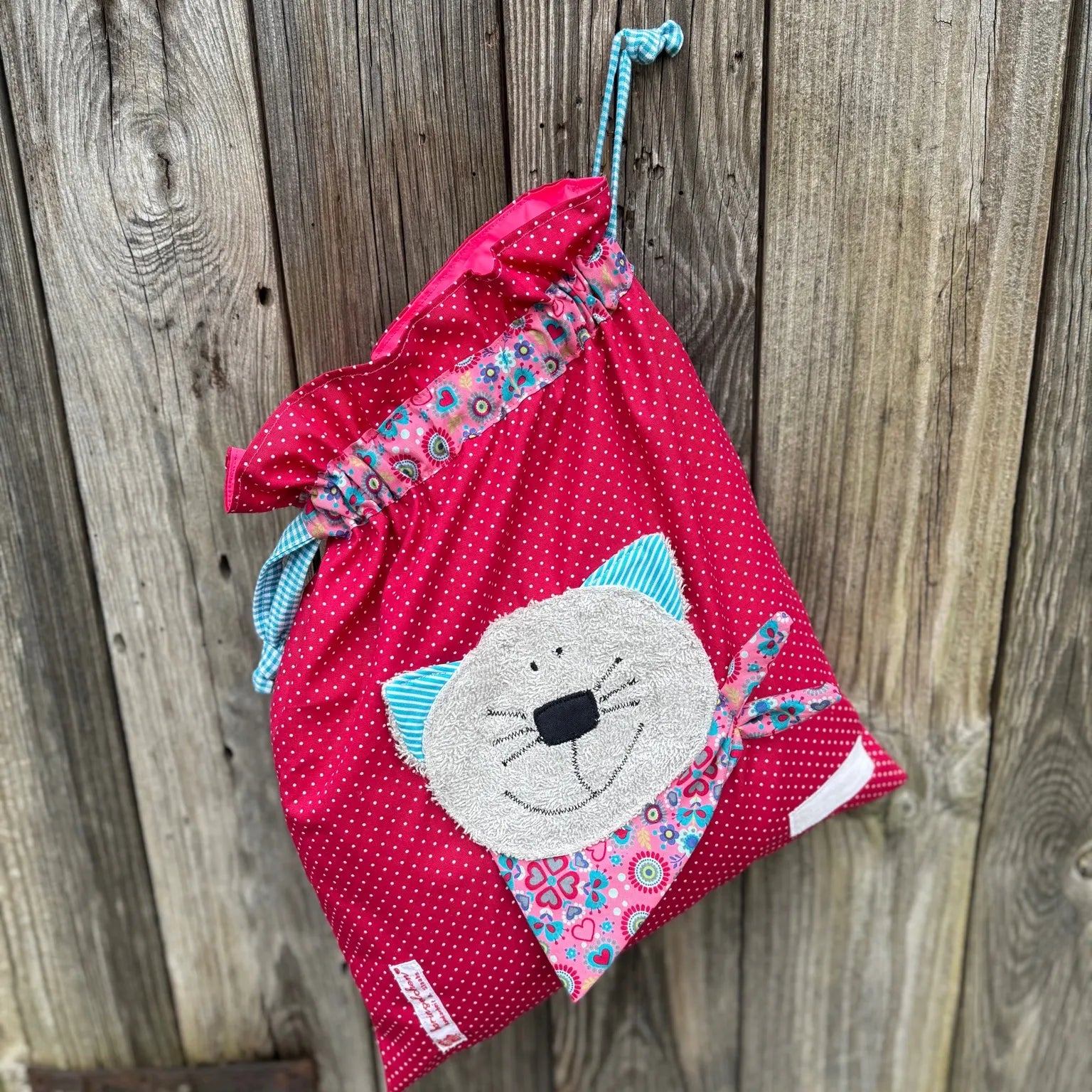 Pinker Wetbag mit weißen Punkten, verziert mit einer bunten Stoffapplikation einer Katze aus grauem Frottee .DerBeutel hat einen floralen Tunnelzug und türkise Kordeln, aufgenommen vor rustikalem Holzhintergrund.