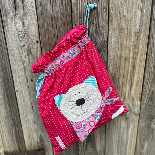 Pinker Wetbag mit weißen Punkten, verziert mit einer bunten Stoffapplikation einer Katze aus grauem Frottee .DerBeutel hat einen floralen Tunnelzug und türkise Kordeln, aufgenommen vor rustikalem Holzhintergrund.