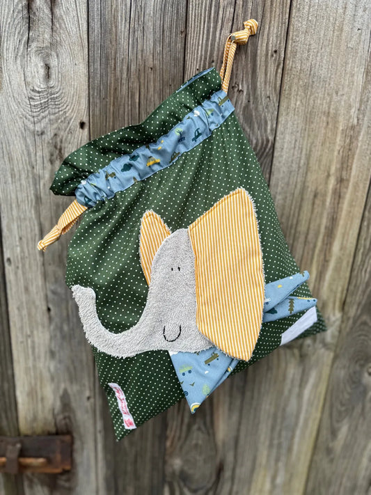 Dunkelgrüner Wetbag mit weißen Punkten, verziert mit einer bunten Stoffapplikation einem Elefant .DerBeutel hat einen blauen Tunnelzug aus  und senfgelben Kordeln, aufgenommen vor rustikalem Holzhintergrund.