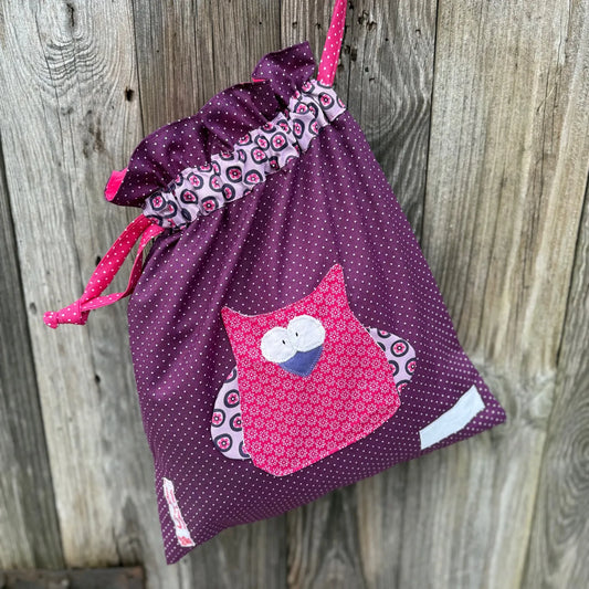Dunkellila Wetbag mit weißen Punkten, verziert mit einer bunten Stoffapplikation einer Eule aus pink gepunkteten Stoff.DerBeutel hat einen floralen Tunnelzug und pinke Kordeln, aufgenommen vor rustikalem Holzhintergrund.