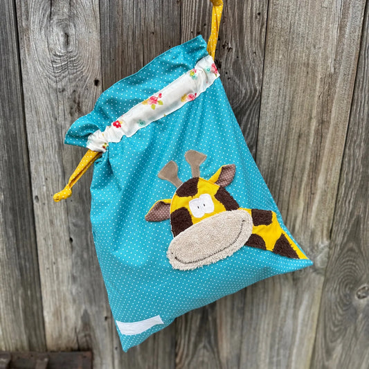Türkiser Wetbag mit weißen Punkten, verziert mit einer bunten Stoffapplikation einer Giraffe .DerBeutel hat einen floralen Tunnelzug und gelbe Kordeln, aufgenommen vor rustikalem Holzhintergrund.