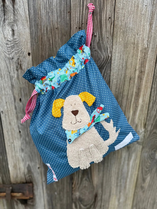 Jeansblauer Wetbag mit weißen Punkten, verziert mit einer bunten Stoffapplikation einer Hund mit  aus hellbraunem Frottee .DerBeutel hat einen Tunnelzug mit
 Fahrzeugprint und rot karierte Kordeln, aufgenommen vor rustikalem Holzhintergrund.